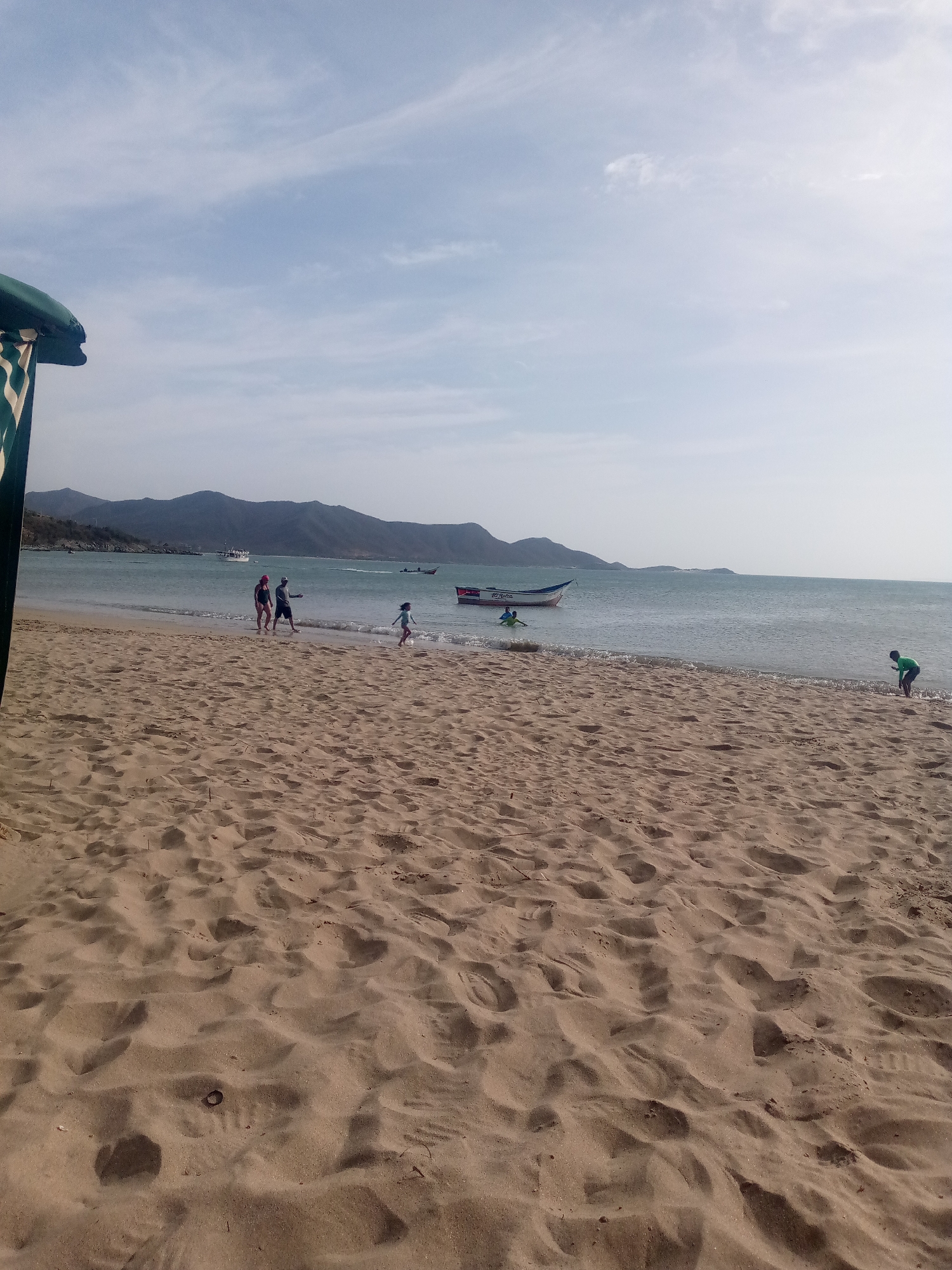 The image shows a beach scene in Juan Griego, Venezuela. Several people are present: a couple walking along the shore, a child running, and two children playing near a boat. A green-clad person is near the water at the far right. Boats are visible in the water. The background includes mountains and a partly cloudy sky. The foreground is dominated by sandy beach.