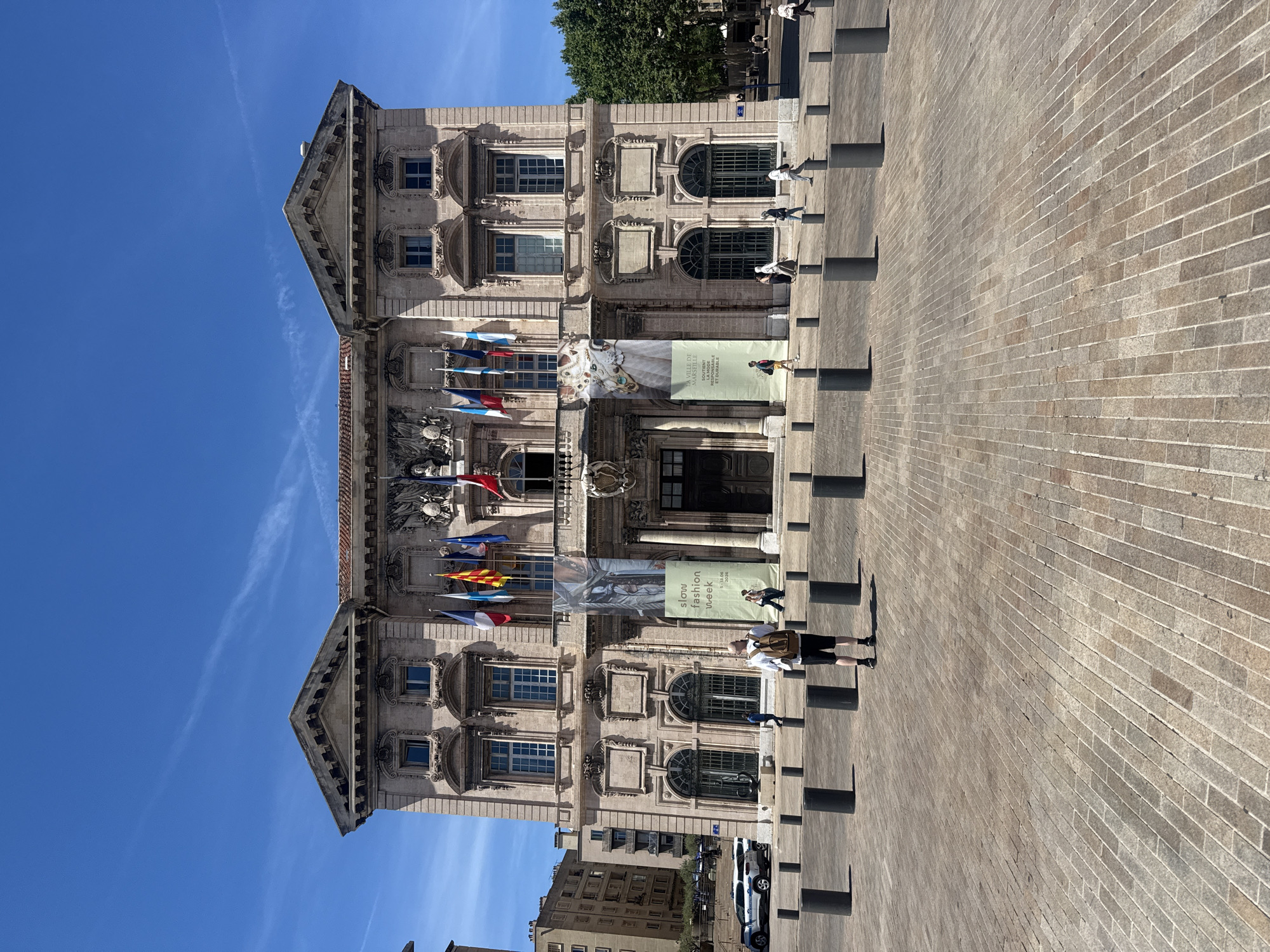 A wide shot captures the facade of a large, ornate stone building under a clear blue sky. Flags fly from the building's upper level. Two large banners hang from the facade, one with text and an image of fabric, the other with text and an image of a jewel-encrusted necklace. In front of the building, a paved plaza is dotted with black bollards. Several people are present in the plaza, including a person with a backpack walking away from the camera, another person standing near a police car, and a few others walking in the distance. The architecture of the building is classical, with arched windows and decorative elements. The overall scene suggests an urban environment with historical architecture and public space.