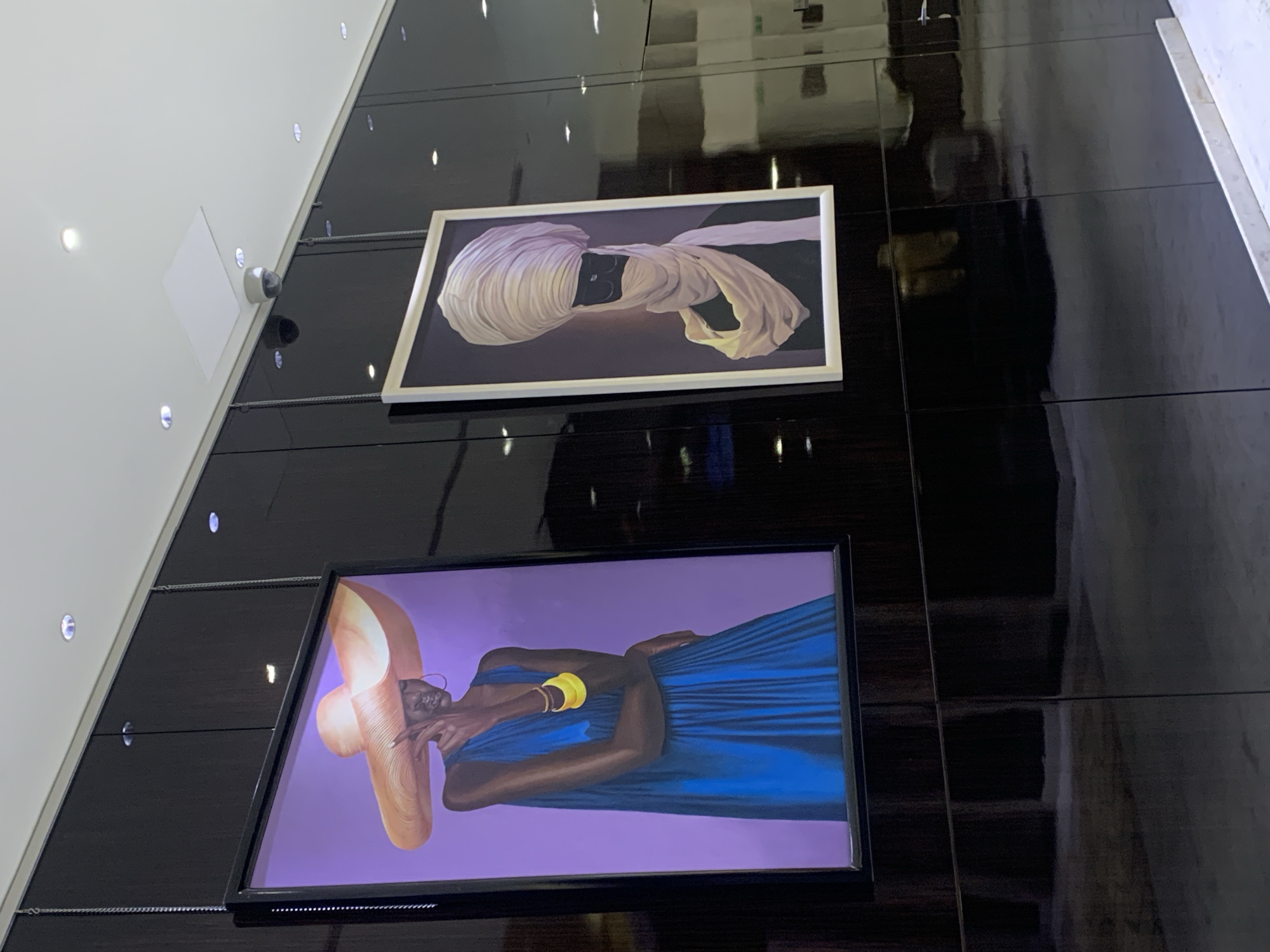 The image shows two framed artworks hanging on a glossy, dark-colored wall in what appears to be an interior space in Dar es-Salaam, Tanzania. The lower artwork depicts a woman wearing a large hat and a blue dress. She is wearing a yellow arm band. The upper artwork depicts a person with their face covered in cloth. A security camera is visible near the ceiling. The wall is made of glossy, dark tiles that appear to reflect the light from the surrounding environment. The scene suggests a modern or contemporary art gallery or upscale retail space. The lighting is bright.