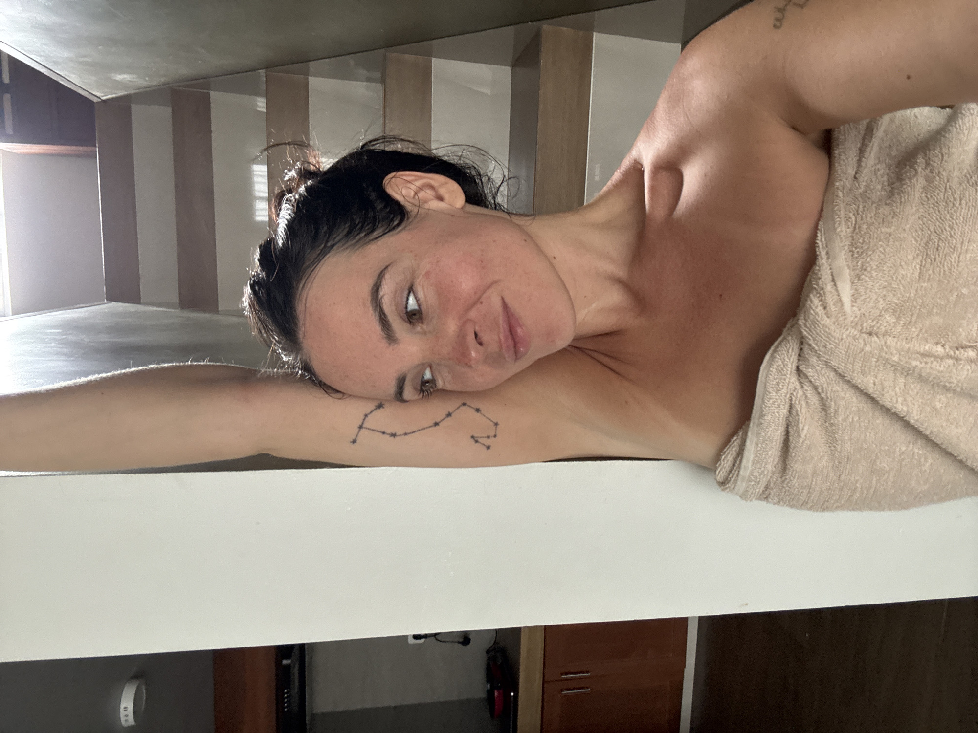 A woman with dark hair pulled back is photographed from the chest up, wrapped in a light brown towel. She stands next to a white wall, with her right arm raised and leaning against it. Her left arm is bent at the elbow, and her left hand is not visible. She has a constellation tattoo on her upper left arm. Her face is turned to the right, and she is looking away from the camera with a slight smile. The background shows a staircase with wooden steps and white risers, and beyond that, a bright light source from above. To the left of the woman, a portion of a kitchen is visible, with wooden cabinets and a countertop. The setting appears to be an indoor residential space in North Kuta, Indonesia.