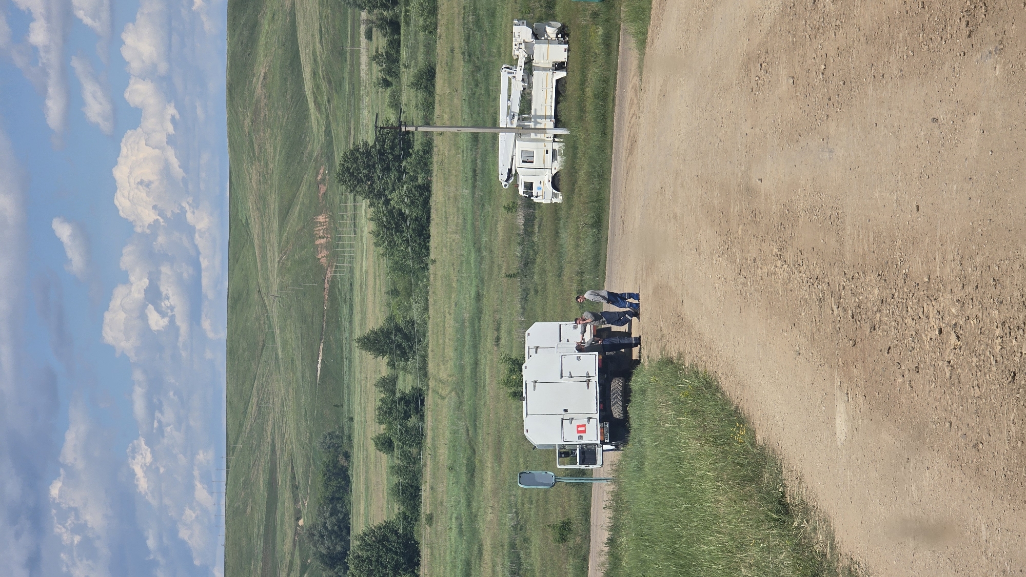 In Ekaterinovka, Russia, on a dirt road bordered by tall green grass, three men in work clothes stand near a white utility truck with its rear door open. One man leans against the truck, while another gestures with his hand, and the third stands with his arms crossed. A second white truck with a large crane is parked further down the road, partially in a grassy field. In the background, rolling green hills meet a cloudy blue sky.