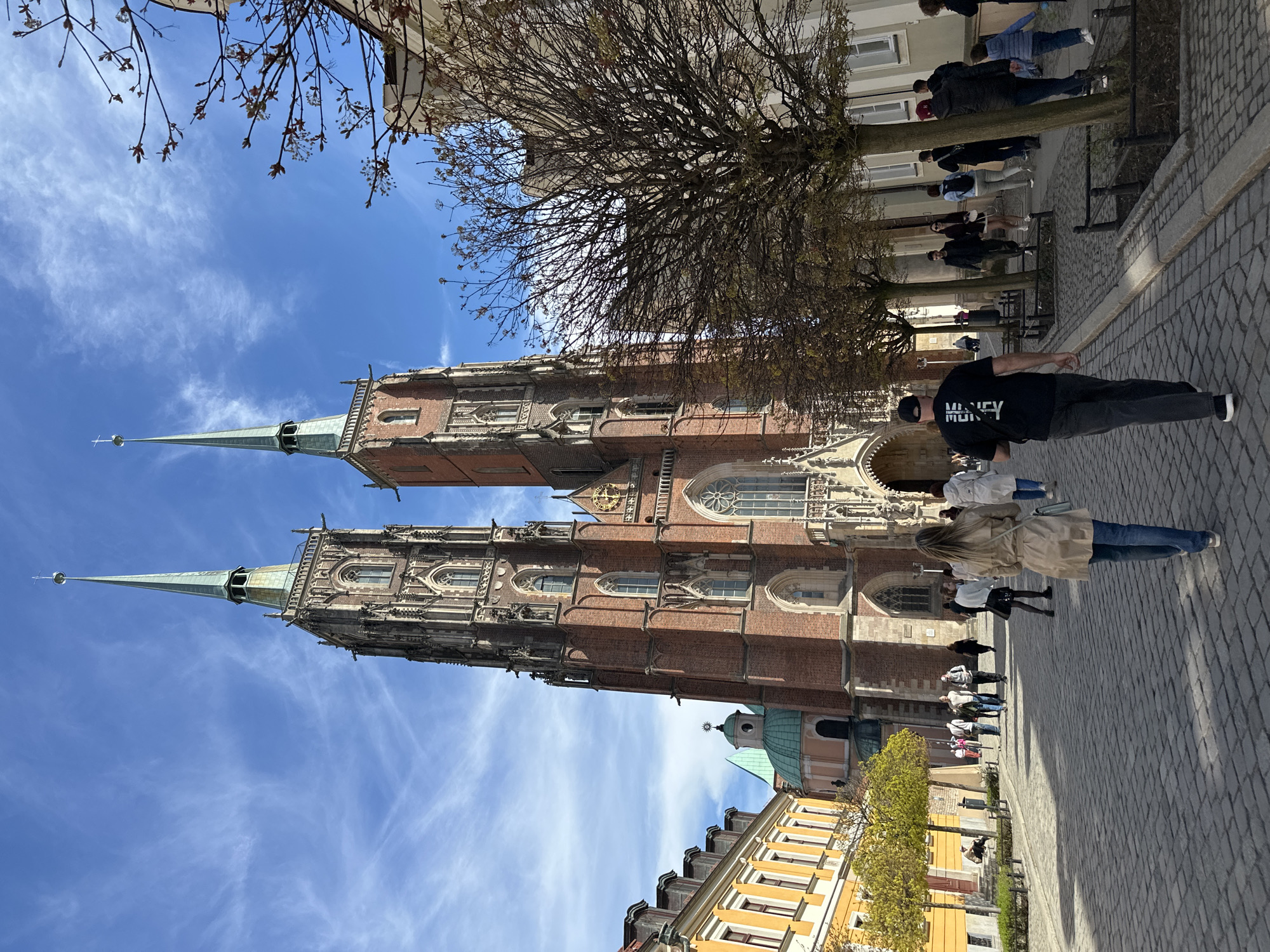 The image depicts a view of the Collegiate Church of the Holy Cross and St. Bartholomew, located on Piasek Island in Wrocław, Poland. The twin-spired Gothic church dominates the frame, with its brick façade and intricate architectural details. A clear blue sky with light clouds is visible behind the church spires. In the foreground, a cobblestone street is filled with pedestrians walking. A bare tree stands to the side, partially obscuring the view of the church. Below the church, various buildings are visible, including structures with yellow facades and green-domed roofs. People are casually strolling along the cobblestone street. The image captures a vibrant urban scene with a blend of historical architecture and modern pedestrian activity.