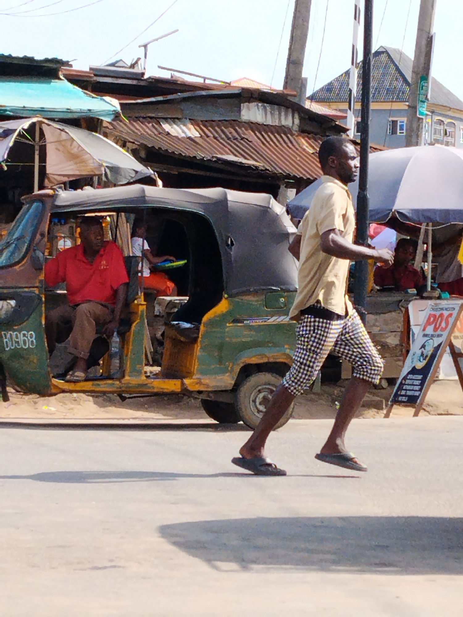 A medium shot reveals a street scene in Aba, Nigeria. A man in a light tan short-sleeved shirt and black and white checkered knee-length shorts is running across the street wearing dark flip-flops. To the left of the man is an old auto-rickshaw painted primarily green with the number "B0968" displayed. Inside the auto-rickshaw, a man is sitting, wearing a red shirt and brown pants. Behind the auto-rickshaw are various stalls with awnings. To the right of the man is a sign that reads "POS." Behind the sign, a person is sitting under an umbrella. Power lines and poles are visible in the background, as well as buildings with corrugated iron roofs. The road is light grey and appears to be paved.