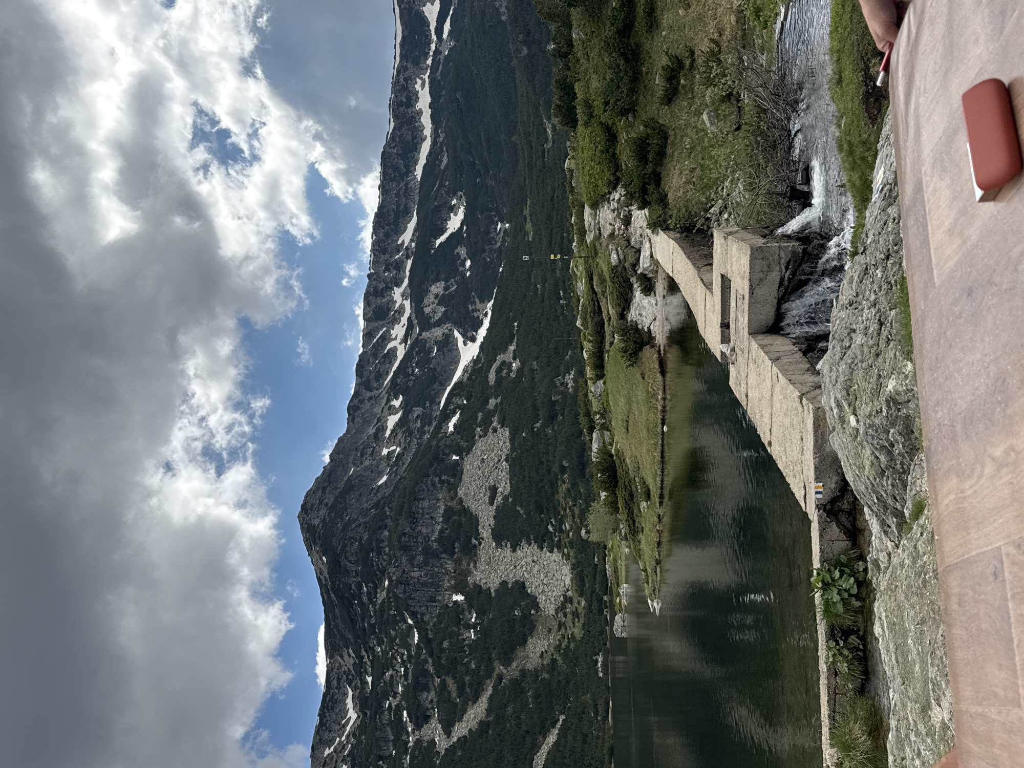The image depicts a scenic mountain lake with a dam structure. The dam, constructed of concrete blocks, runs across a narrow channel where water flows, creating a small waterfall. The lake's surface is calm and reflects the surrounding landscape. On the far side of the lake, a lush green mountainside rises, dotted with trees and patches of snow, culminating in a rocky summit. The sky is partly cloudy with dramatic formations of white and grey clouds against a blue backdrop. In the foreground, a wooden surface, possibly a table, is visible, with a red object and a hand holding a red object resting on it. The scene suggests an outdoor recreational area. The specific location is Avramovo, Bulgaria.