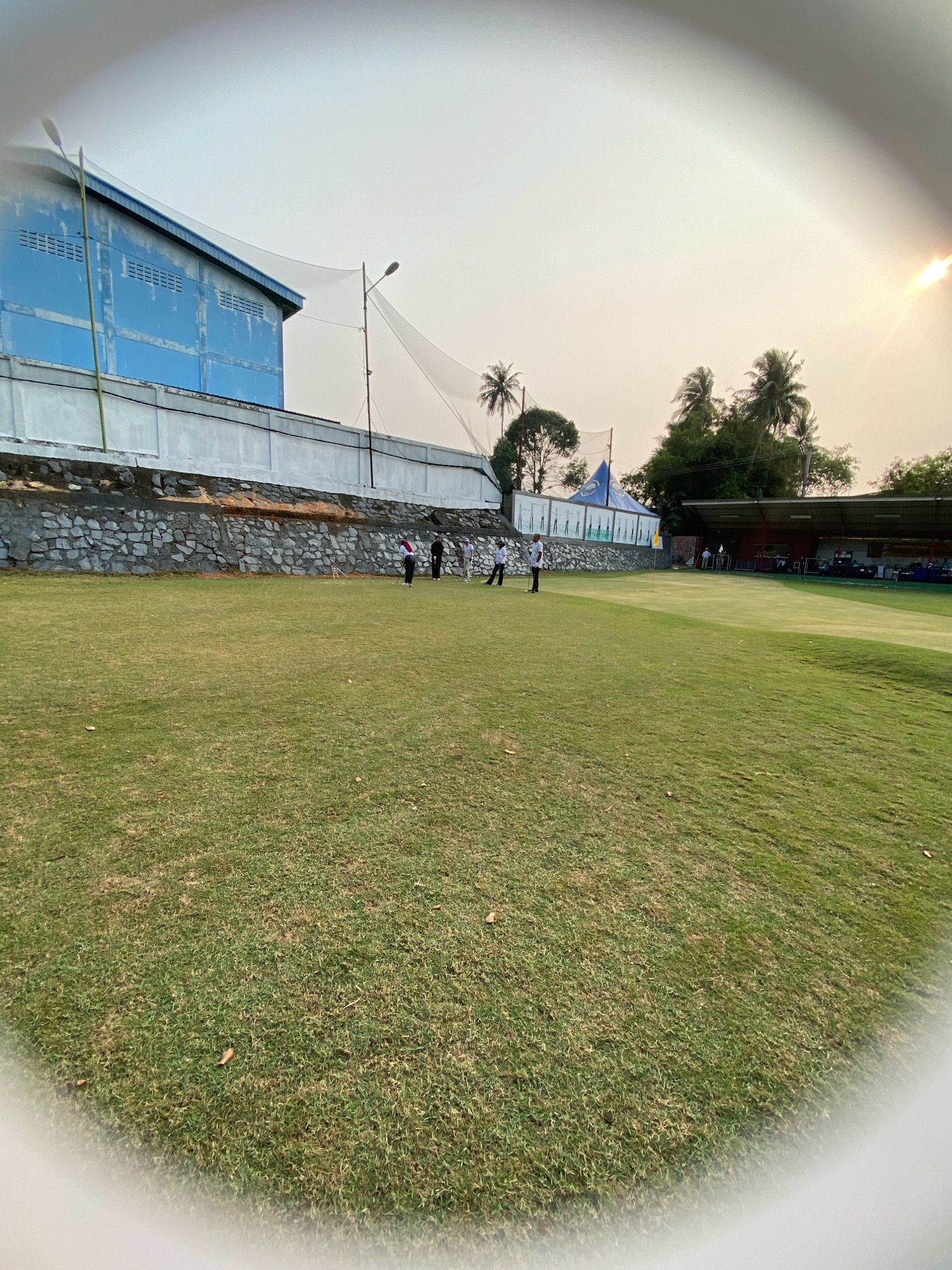 The image depicts a golf course in Tanjung Pinang, Indonesia, with a large, light blue industrial building in the background. The building has a white trim and several rows of horizontal vents. A white wall with a stone foundation runs along the perimeter of the building. A large net, presumably to protect from golf balls, is suspended behind the building. Coconut trees and other foliage are visible beyond the building and net. The foreground is dominated by a lush green golf course, with a few golfers visible in the middle ground. One golfer in a white shirt and dark pants is positioned near a yellow flagstick on the green. The sky is bright and hazy, suggesting a warm day, with the sun glinting in the upper right corner. The image has a fisheye lens effect, distorting the edges and creating a curved appearance. A shaded structure with stadium seating is visible on the right side of the golf course.