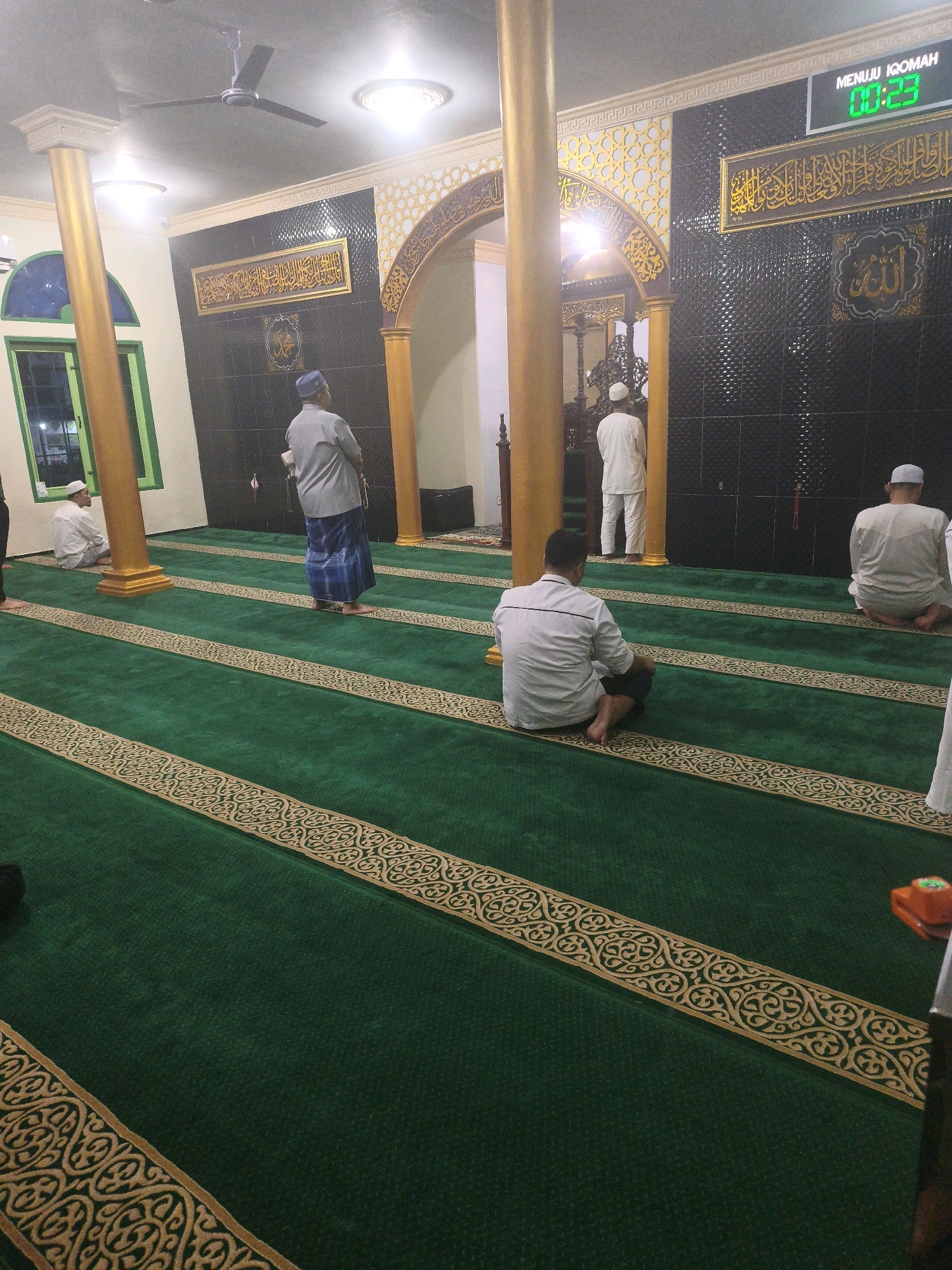 The image depicts the interior of a mosque in Samarinda, Indonesia. The mosque features a green carpet with gold decorative borders, and several men are present, engaged in prayer or waiting. In the foreground, a man in a white shirt is seated on the carpet, facing away from the camera. Further back, a man in a grey shirt and blue sarong stands facing towards an arched doorway. Another man in a white robe stands in the arched doorway, facing away. On the right side of the image, a man in a white shirt is seated with his back to the camera. The walls are decorated with intricate gold calligraphy and a tile mosaic. A digital clock displaying "00:23" and the text "MENUJU IQOMAH" is visible on the upper right wall, indicating it's time for prayer. The lighting is provided by ceiling lights and natural light coming through a green-framed window on the left. A ceiling fan is visible.