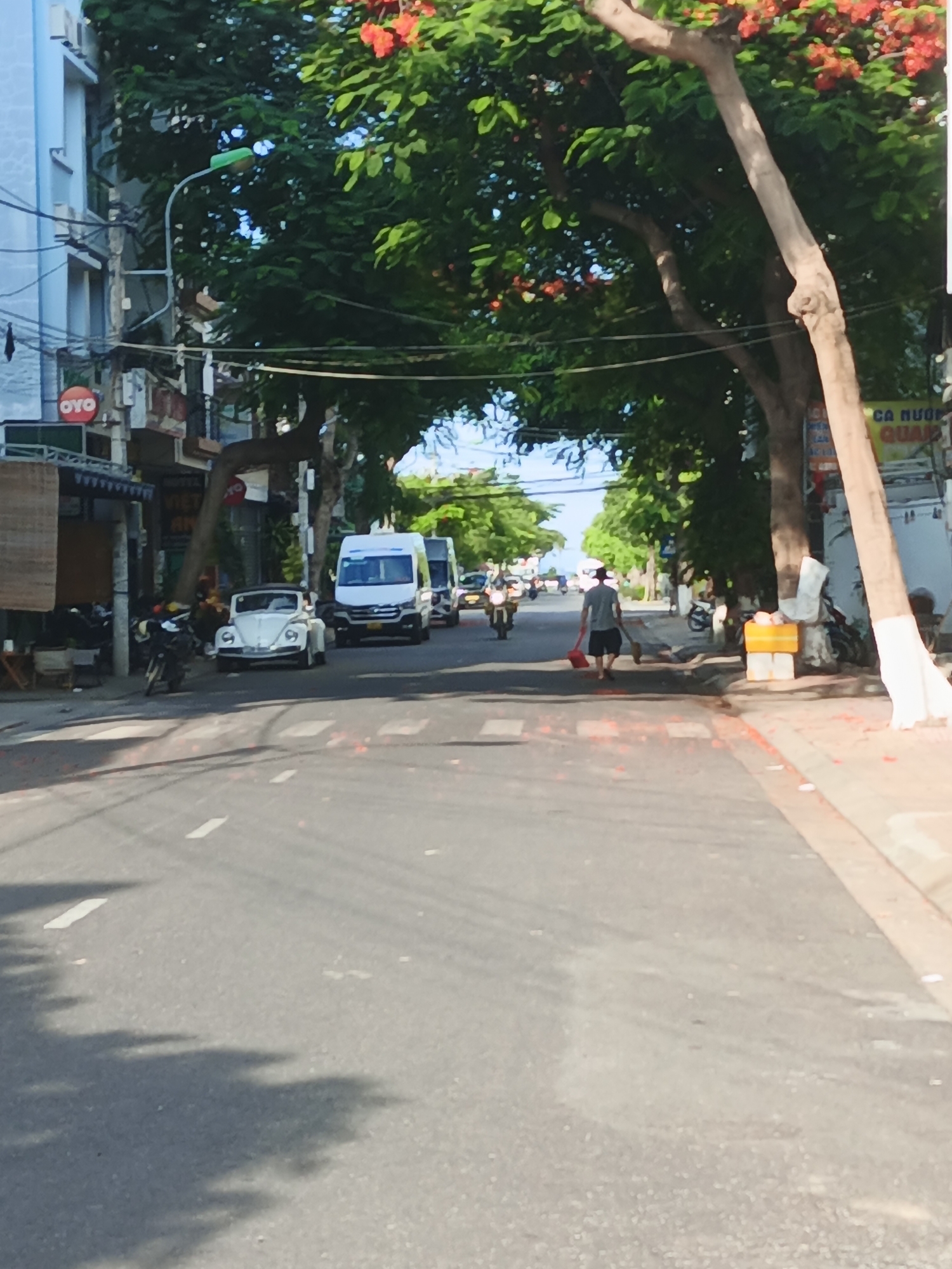 In the city of Nha Trang, Vietnam, on a street lined with trees and buildings, a man wearing a hat and shorts walks across the road, holding a red broom and a dustpan. The street is paved and has white lane markings. A white vintage Volkswagen Beetle is parked on the left side of the road, with a white van and other vehicles further down the street. A motorcycle is parked in front of a building. The buildings on the left appear to be commercial establishments, with signs visible. Red flowers are blooming in the trees overhead, casting shadows on the road. There are yellow bins on the right side of the street. The scene depicts a typical day on a city street in Vietnam.