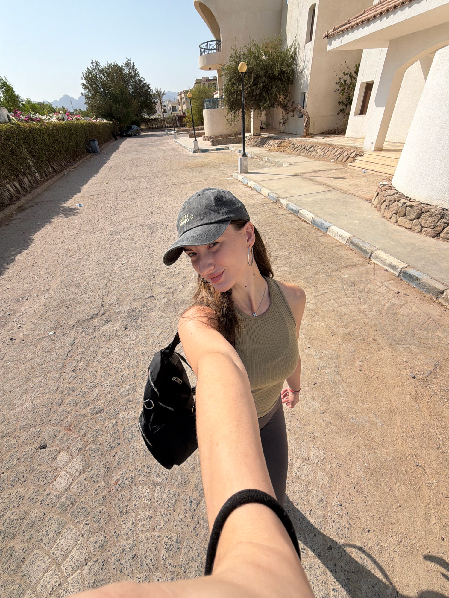 A woman is taking a selfie while walking on a paved street in Sharm El Sheikh, Egypt. The woman is wearing a dark baseball cap, a green tank top, and grey leggings. She has a black bag slung over her shoulder. The background shows buildings with white facades, a hedge of flowering plants, and a clear blue sky. The street is paved with asphalt and lined with sidewalks and decorative stonework. The lighting suggests it is daytime, and the sun is casting shadows.