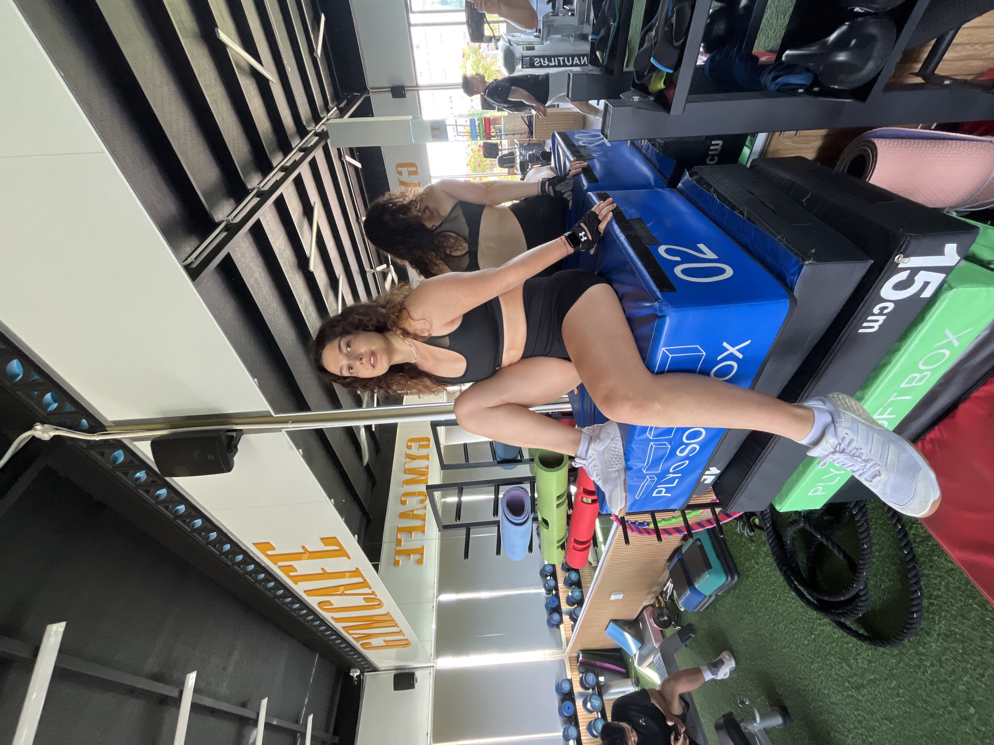 The image captures a woman in athletic wear sitting atop a stack of plyometric boxes in a gym. She is positioned on the left side of the frame, facing the camera, with her legs bent and one foot resting on the top blue box. Her right hand is on her knee, and her left hand is resting on the box. She has curly dark hair and is wearing a black sports bra and black shorts. Her white athletic shoes are visible.
The gym environment is evident with various exercise equipment in the background. To the left, there are rows of dumbbells and exercise mats. A mirror reflects the space, showing a "GYMCAFE" sign in large orange lettering. In the distance, other gym-goers are visible, one of whom is looking at their phone. The floor appears to be artificial turf.
The image is described as being located in the city of Phan Thiết, Vietnam.