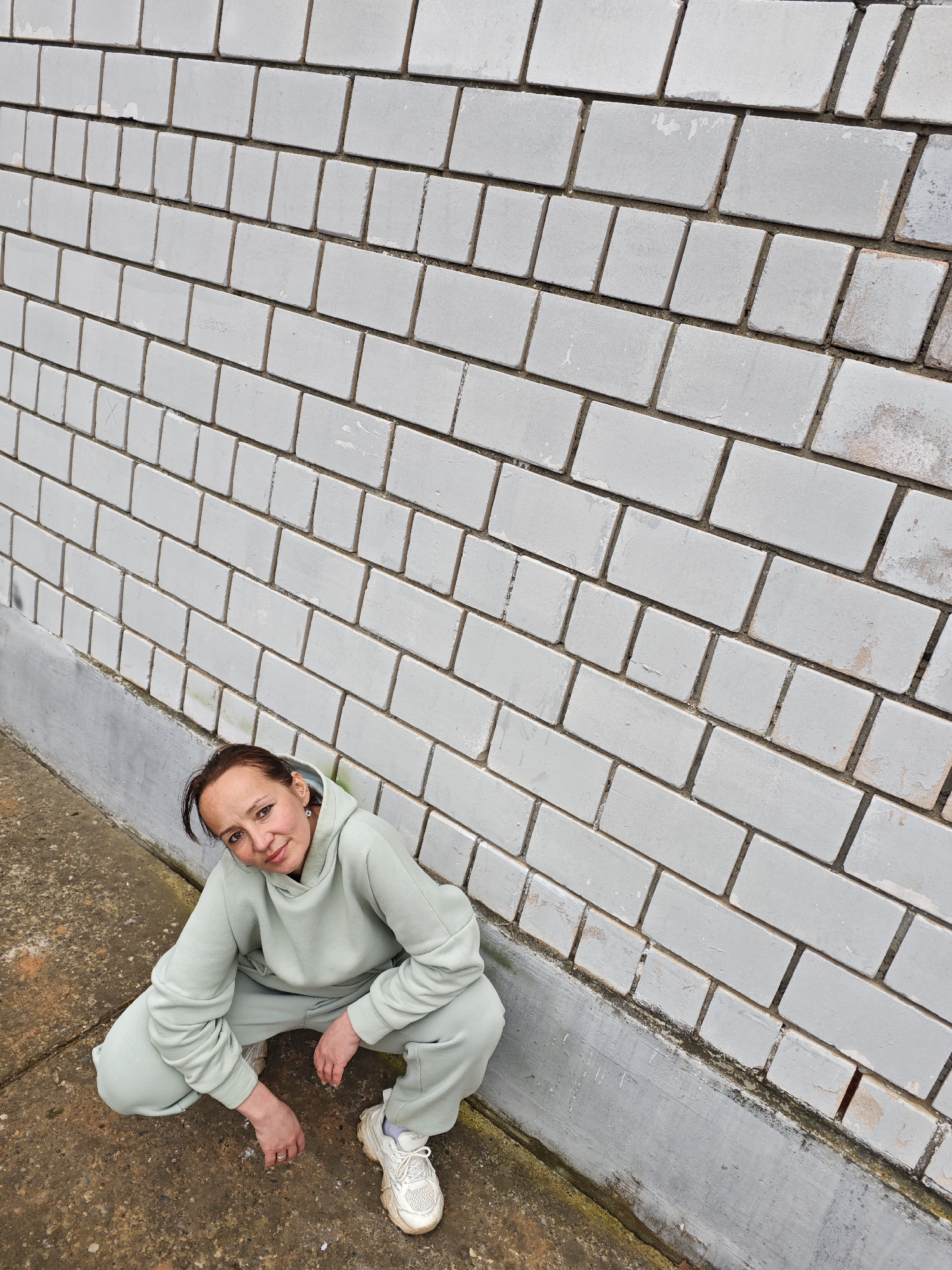 A woman is crouching in front of a brick wall in Марковский, Russia. The wall is light grey, made of rectangular bricks with darker grout. The woman wears a matching light green hoodie and sweatpants and white sneakers. She is looking at the camera, her hands resting on the ground. The ground is a mix of grey and brown.