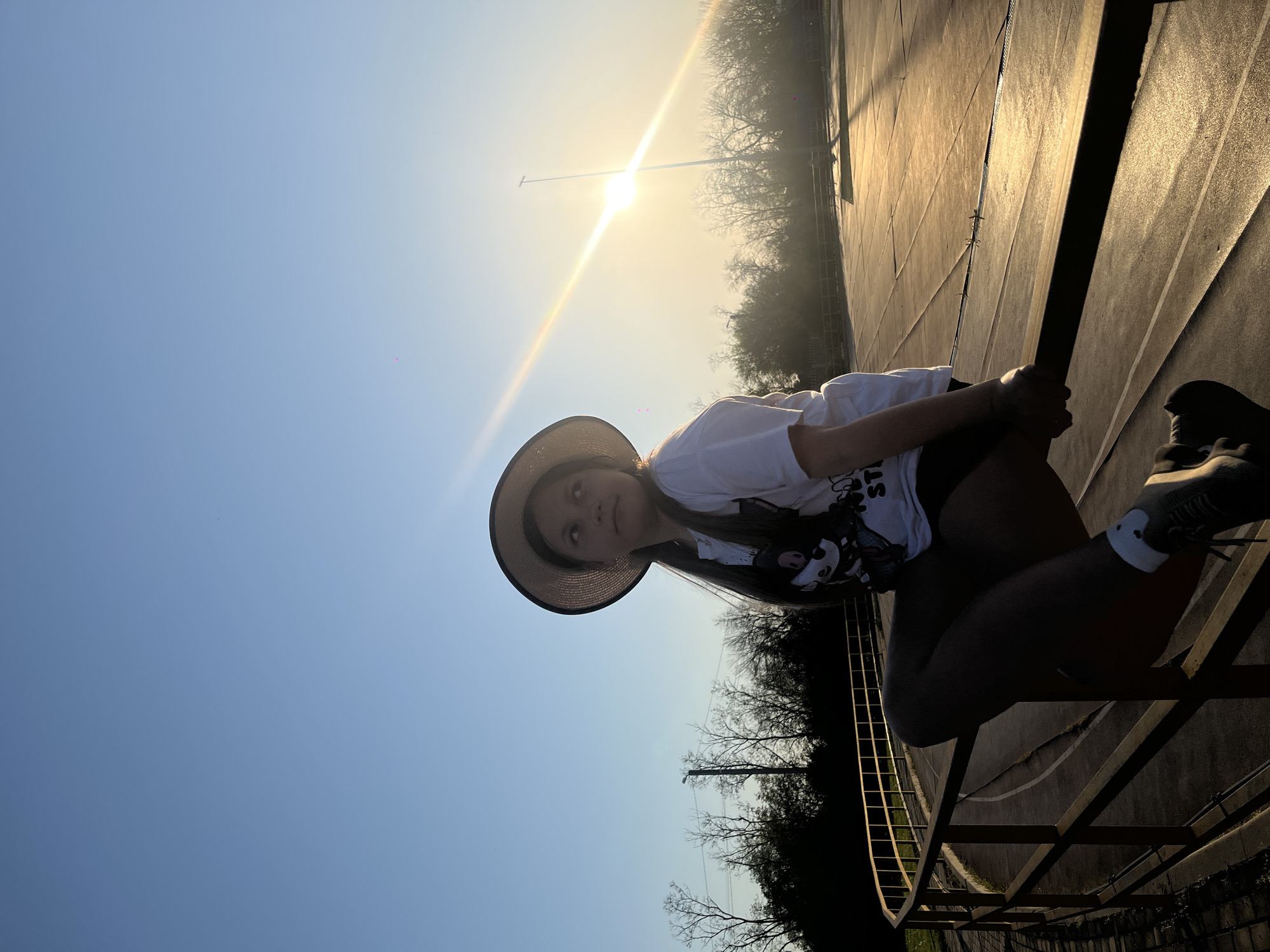 A young woman wearing a white t-shirt, dark shorts, and a straw hat is sitting on a metal structure. She is leaning against a railing, holding onto it with her hands. The background features a bright sky, trees, and a paved surface. The location appears to be outdoors, possibly a park or recreational area in Novopokrovskaya, Russia.