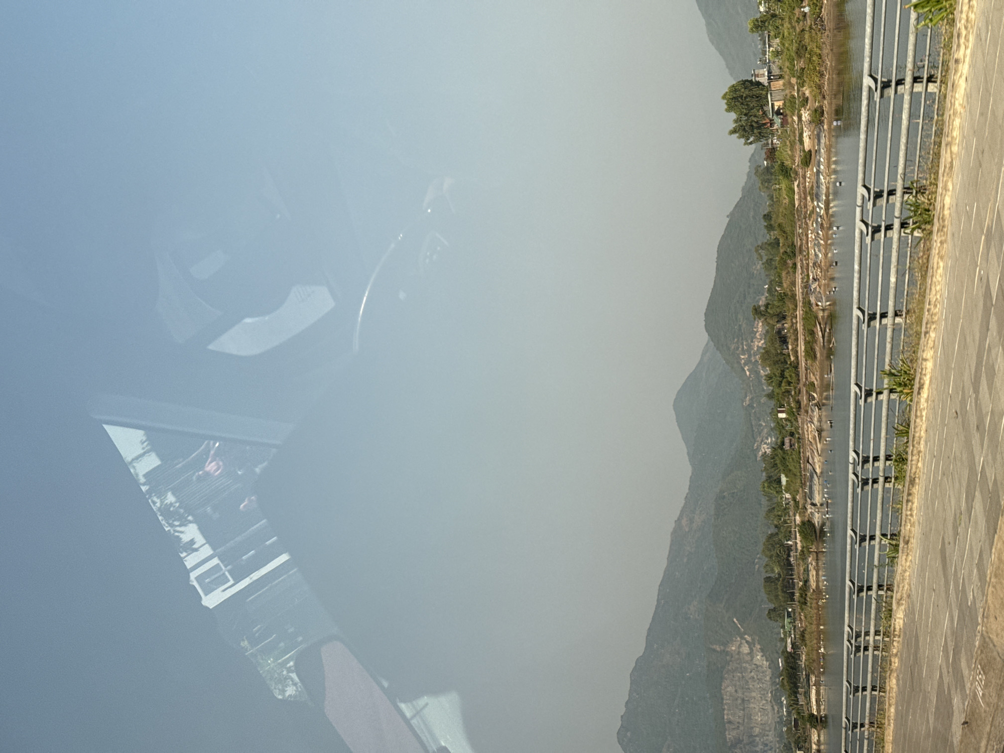 The photograph, taken from the inside of a car, presents a view of a waterway in Unknown, Unknown, bordered by a mountain range. The reflections in the car window obscure the left side of the image. The waterway is parallel to a sidewalk and a series of metal railings. Along the waterway, there are scattered trees and buildings. The mountain range forms the backdrop of the scene.