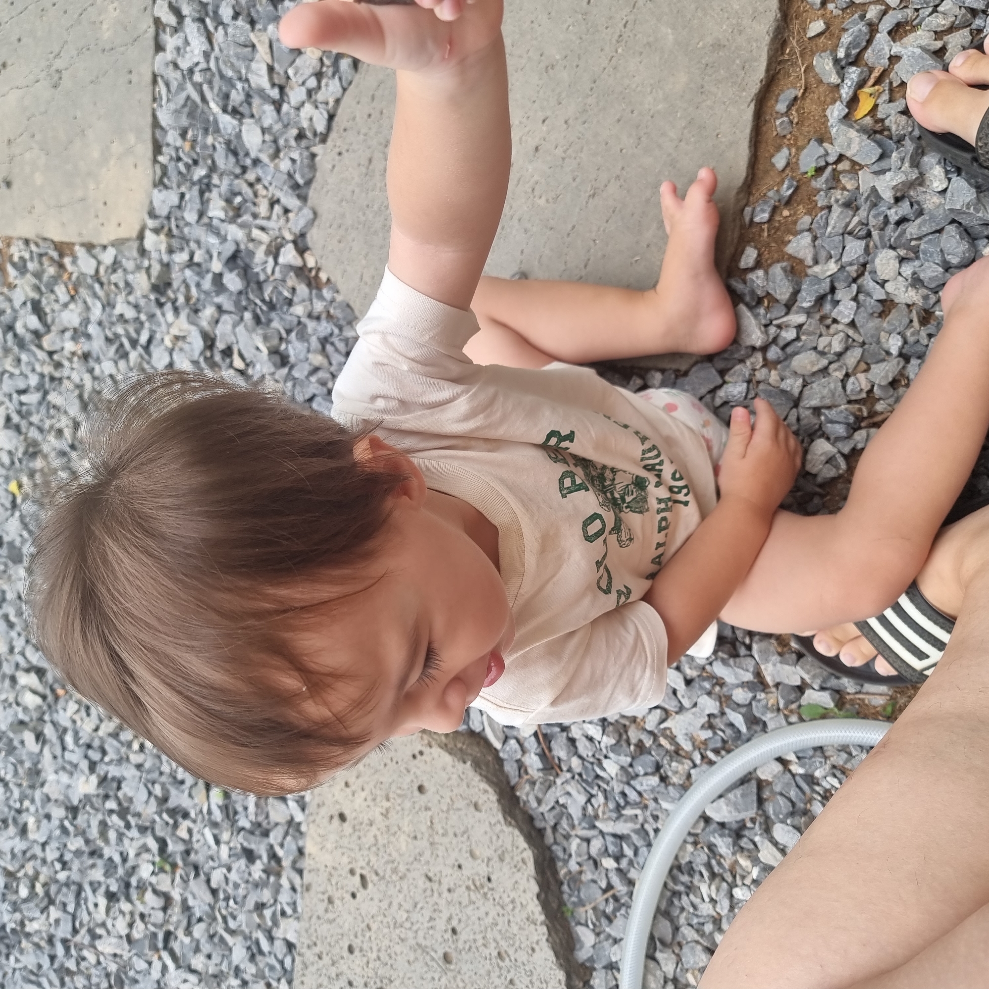A young child with brown hair, wearing a light-colored t-shirt with a graphic print, sits on a surface composed of gravel and large, flat stones. The child is looking down and to their left, with their tongue slightly out. Their right hand is raised, holding a dark object, possibly a twig or a small stone. To the left of the child, a portion of an adult's leg is visible, wearing a black and white striped sandal. A grey hose lies coiled on the gravel to the left of the child. The overall setting appears to be an outdoor area, possibly a yard or patio, with the typical textures and colors of Dunnae-myeon, South Korea.