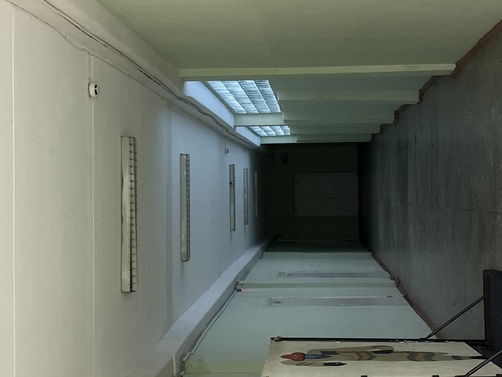 The image depicts a long, narrow corridor or passageway, potentially located in an industrial or institutional setting in Berezniki, Russia.
The corridor's walls are painted in light, muted tones, possibly gray or pale green. The left wall is smooth and features a series of vertical metal or plastic strips affixed to its surface, along with a small white circular object (possibly a sensor or light fixture). The right wall appears rougher, with a textured, possibly concrete, surface and a stepped or uneven ceiling line.
The ceiling incorporates translucent, possibly glass block, skylights that allow natural light to filter into the space. This light diminishes towards the far end of the corridor, creating a sense of depth and perspective.
The floor is marked with rectangular shapes, likely indicating tiles or concrete slabs. In the foreground, a portion of an object, possibly a rug or tapestry, is visible.
The overall impression is of a utilitarian and possibly aging space, lacking decorative elements and primarily designed for function rather than aesthetic appeal. The lack of people and specific context leaves the purpose of the corridor ambiguous.