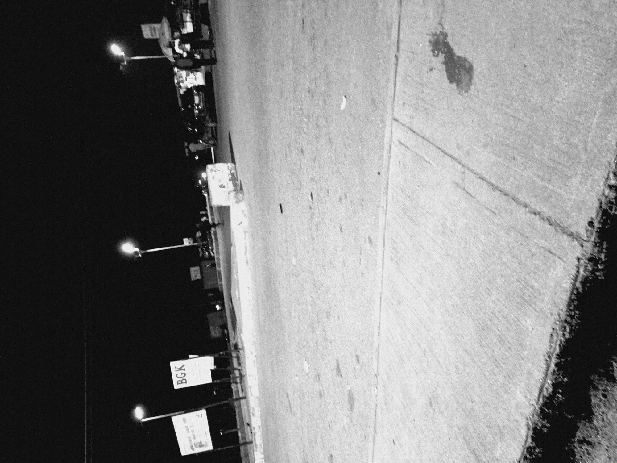 The image depicts a street scene at night in Bukuru, Nigeria. The foreground is dominated by a concrete surface, possibly a sidewalk or road, with a distinct dark edge. In the midground, a street with a tarmac surface is visible. Several streetlights illuminate the scene, casting a harsh glow on the surroundings.
Two prominent signs are present: one on the left, largely obscured by darkness and motion blur, and another in the center displaying the letters "BGK" in a block font. Beyond these, the scene shows a busy area with what appear to be stalls or shops, some with awnings. There are several people visible, some standing near the stalls and others walking. A few vehicles are also present, including a car parked on the right side of the road. The overall atmosphere is that of an active street or market at night, with artificial lighting providing the primary illumination.