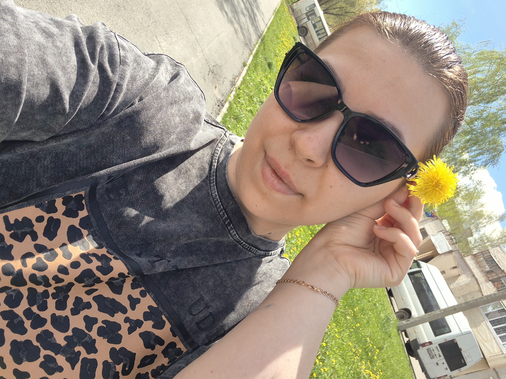 A woman is holding a yellow dandelion near her ear, with a white van and building in the background. She wears black sunglasses, a gray acid-wash long-sleeved shirt with a leopard-print pattern underneath, and a gold bracelet on her left wrist. The scene is outdoors on a sunny day with grass and yellow dandelions visible. The buildings and vehicles suggest an urban or suburban environment. The location is Tobolsk, Russia.