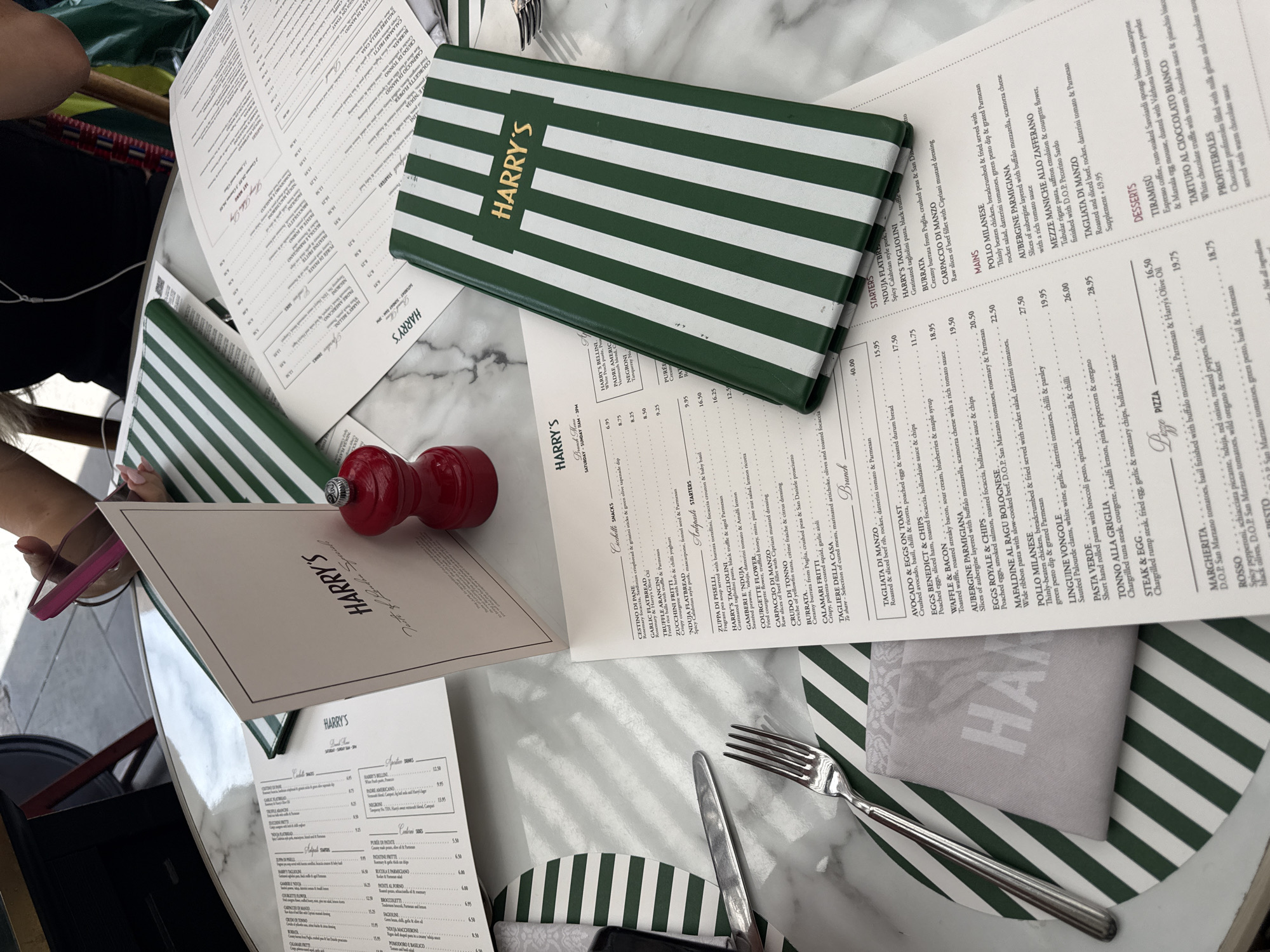 In a well-lit outdoor setting, a marble-topped table displays multiple menus from "Harry's," a restaurant in London, United Kingdom. One menu, partially obscured by a green and white striped cover, is open to a section listing "Starters," "Mains," and "Desserts." To the left, another menu, also with "Harry's" branding, is open to a section labeled "Brunch." A red pepper mill sits prominently on the table between the menus. A silver fork and knife are placed on a patterned charger plate, which has green and white stripes mirroring the menu cover. A folded grey napkin with "Harry's" embossed on it lies on the charger plate. In the background, a person's hands are visible, holding a pink smartphone, and a portion of their upper body is also visible. The overall impression is a casual dining experience at a table set for a meal.