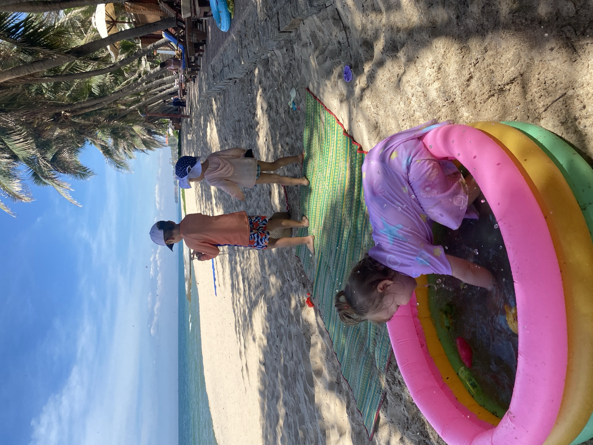 A girl is playing in an inflatable pool on the beach in Phu Thuy Ward, Vietnam. The pool is pink, yellow, and green, and filled with water. The girl is wearing a purple swimsuit and has her hair in pigtails. She is reaching into the pool with her hands, splashing water.
Two children stand on a mat behind the pool. The child on the left is wearing an orange long-sleeved shirt and patterned shorts and a white baseball cap. The child on the right is wearing a light pink hooded shirt, light blue shorts, and a white hat with a blue pattern.
Palm trees line the beach in the background, and lounge chairs are visible under the trees. The ocean is in the distance, with a sandy beach in the foreground. The sky is blue with scattered clouds. The overall scene depicts a sunny day at the beach with children playing.