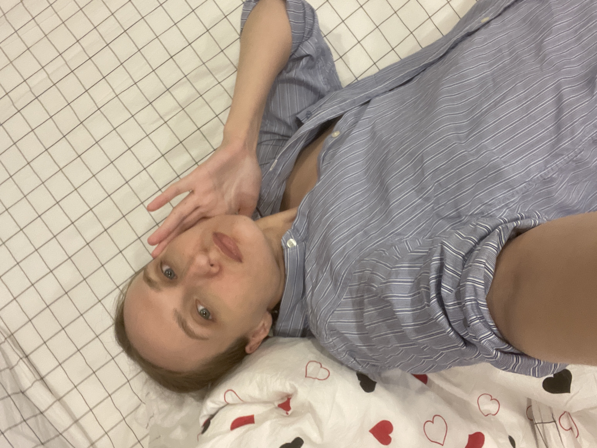 A young woman is depicted lying on her back on a bed indoors, likely in Подшивалово, Russia. She is looking directly at the camera, appearing to take a selfie. Her expression is neutral. She has light-colored hair and eyes, and fair skin. She wears a loose-fitting blue and white striped long-sleeved collared shirt, which is partially unbuttoned at the neckline. Her right arm is raised, with her fingers resting near her face.
The bed is covered with white bedding featuring a light gray grid pattern. Closer to the bottom right of the frame, a pillow or blanket with a pattern of red and black heart outlines, as well as solid red and black hearts, is visible. The lighting is consistent with an indoor setting, and no specific time of day or outdoor weather conditions can be discerned. There is no visible text in the image.