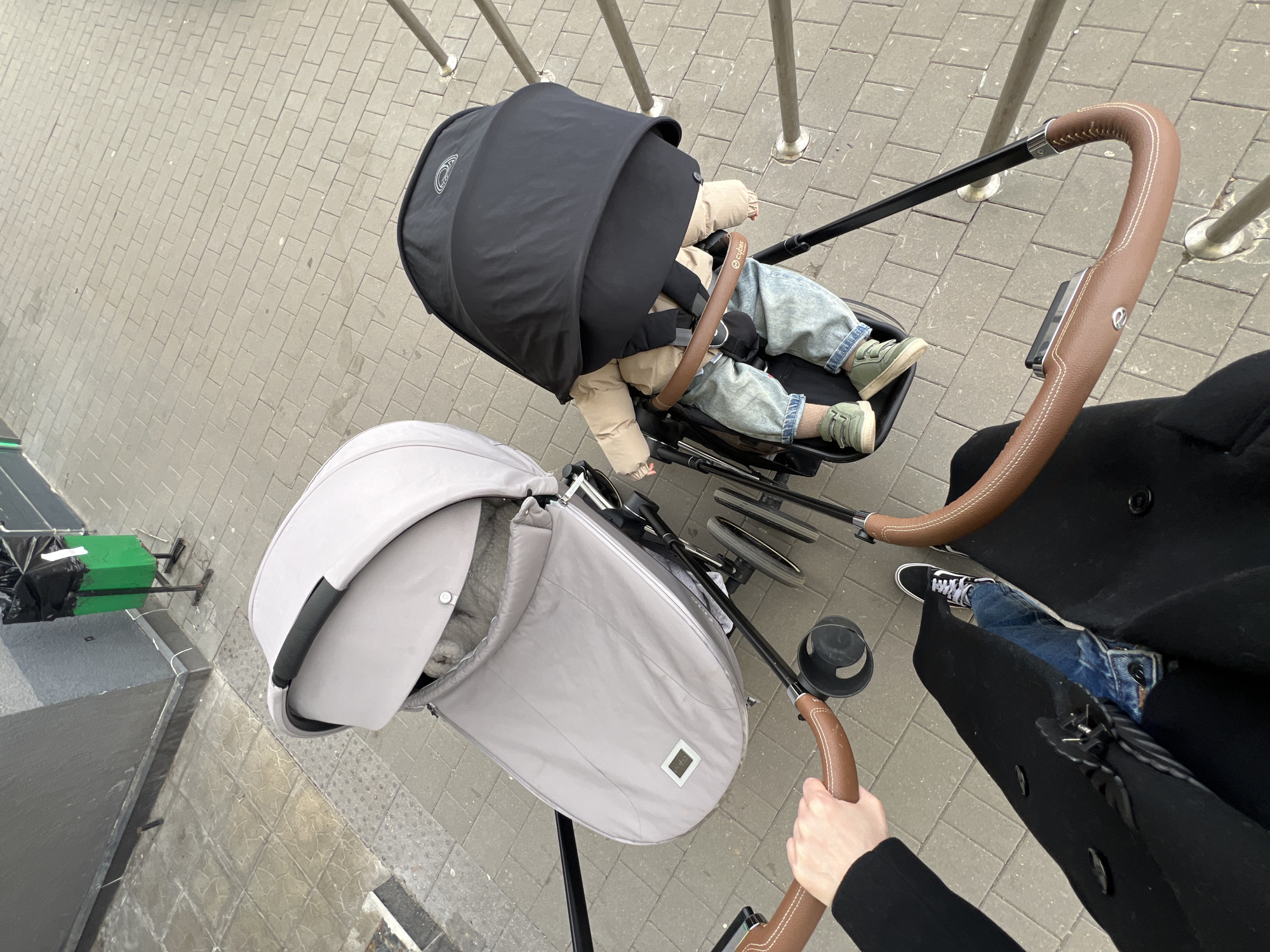 An overhead shot depicts two strollers on a brick-paved surface, likely a street. One stroller, in the upper right, has a dark cover and contains a child wearing jeans and green shoes. The second stroller, in the lower left, is light gray with a partial cover. A person wearing a black coat and blue jeans with black and white sneakers stands to the right, visible from the thighs down, holding the stroller handles. The scene appears to be in Khimki, Russia, based on visual evidence and the availability of the image. Metal railing posts are visible in the background.