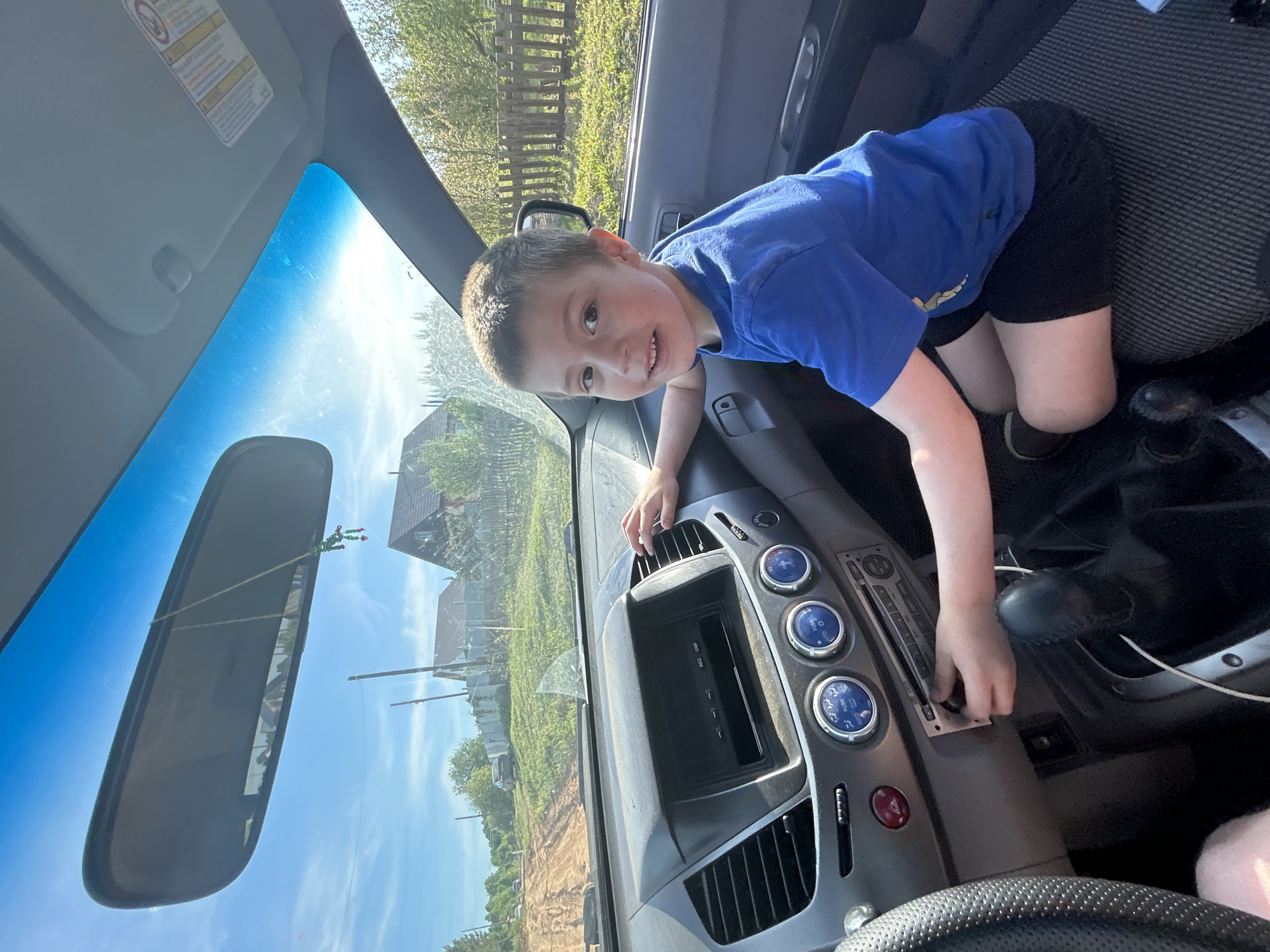 A young boy in a blue t-shirt and black shorts is leaning across the dashboard of a car. He is smiling at the camera and touching the car's controls. Outside the car, a rural landscape is visible. The scene is at Фоки, Russia, which is visible on the car's GPS unit.