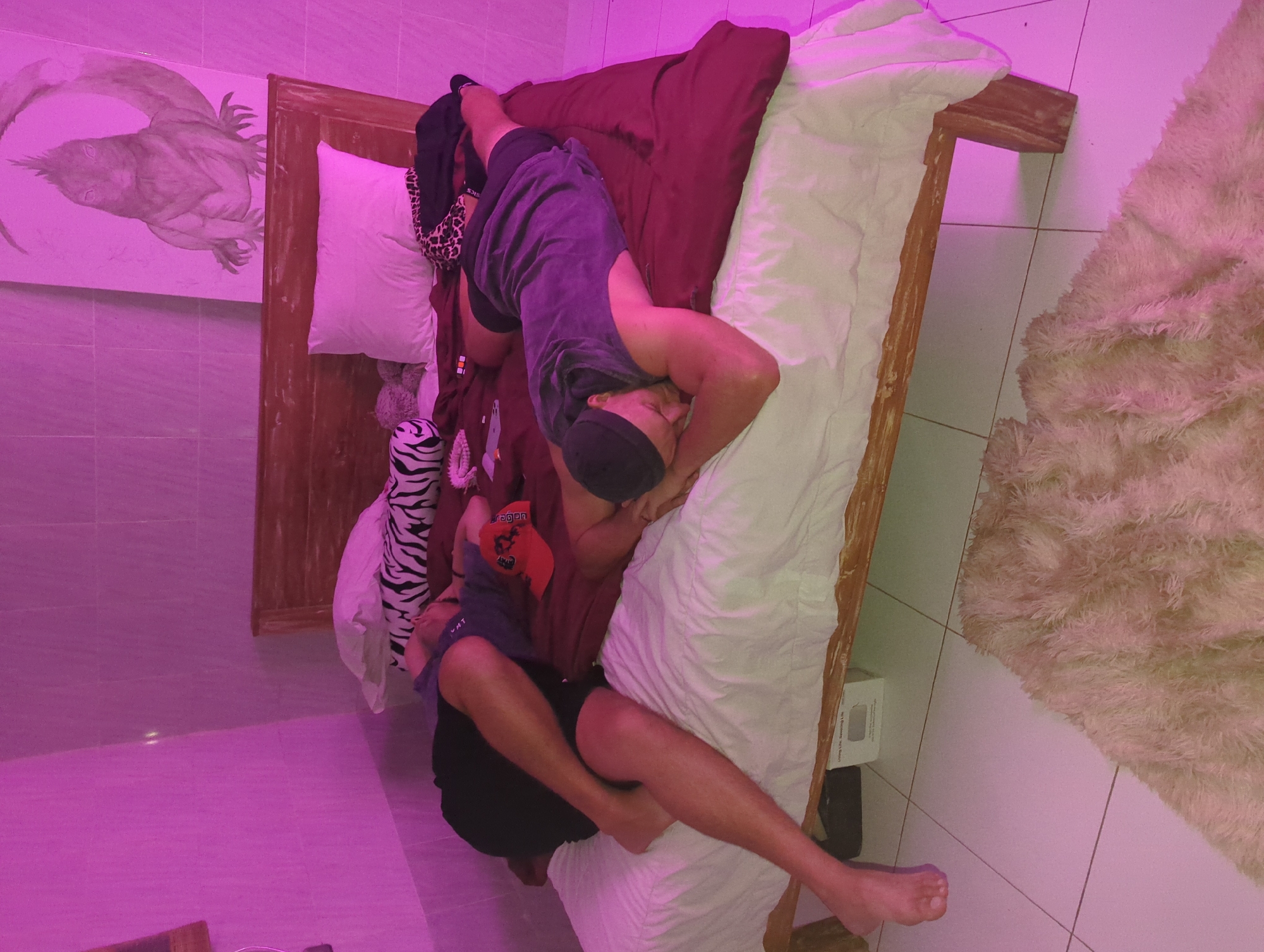 A photograph taken in a room, possibly a bedroom or guest room, in North Kuta, Indonesia, shows two men lying on a wooden-framed bed covered with a red-brown sheet and white bedding. The room is lit with a pinkish hue. The bed is placed against a wall with a mounted painting featuring a stylized animal, possibly a lizard or frog. Pillows of various patterns and colors are scattered on the bed. The floor is tiled, and a light-colored, fluffy rug is partially visible. One man wears a dark sleeveless shirt and dark shorts, and the other wears a similar outfit and a red baseball cap. A product box is visible near the foot of the bed on the floor.