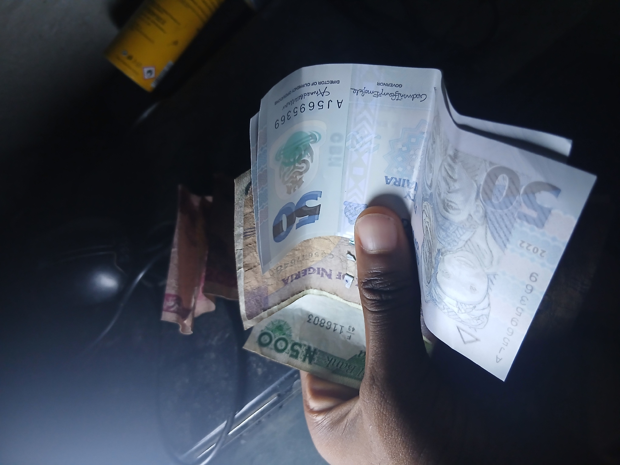 A hand holds Nigerian Naira banknotes. A 500 Naira note is visible at the bottom, partially obscured by the hand. Above it is a bundle of 50 Naira notes. The serial number "AJ5695369" and the signature of "Godwin Ifeanyi Emefiele," the Governor, are visible on the notes. The scene is dimly lit.