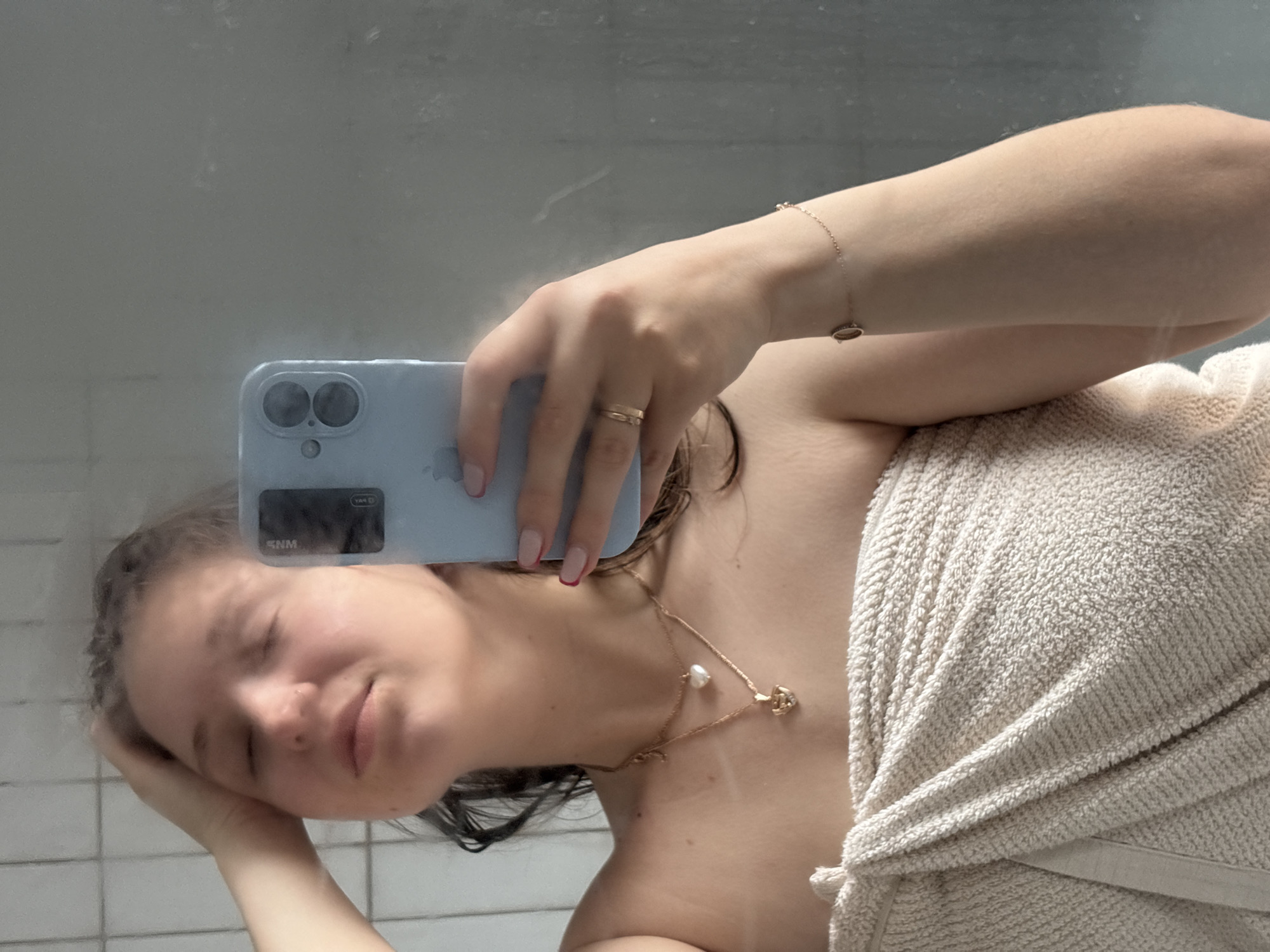 A person with a towel wrapped around their body is taking a selfie in front of a mirror. The person is holding a light blue smartphone, and their face is partially visible. They are wearing jewelry, including a necklace and bracelet. The background shows a tiled wall and a reflection in the mirror. The location of Izhevsk, Russia is not discernible from the image.