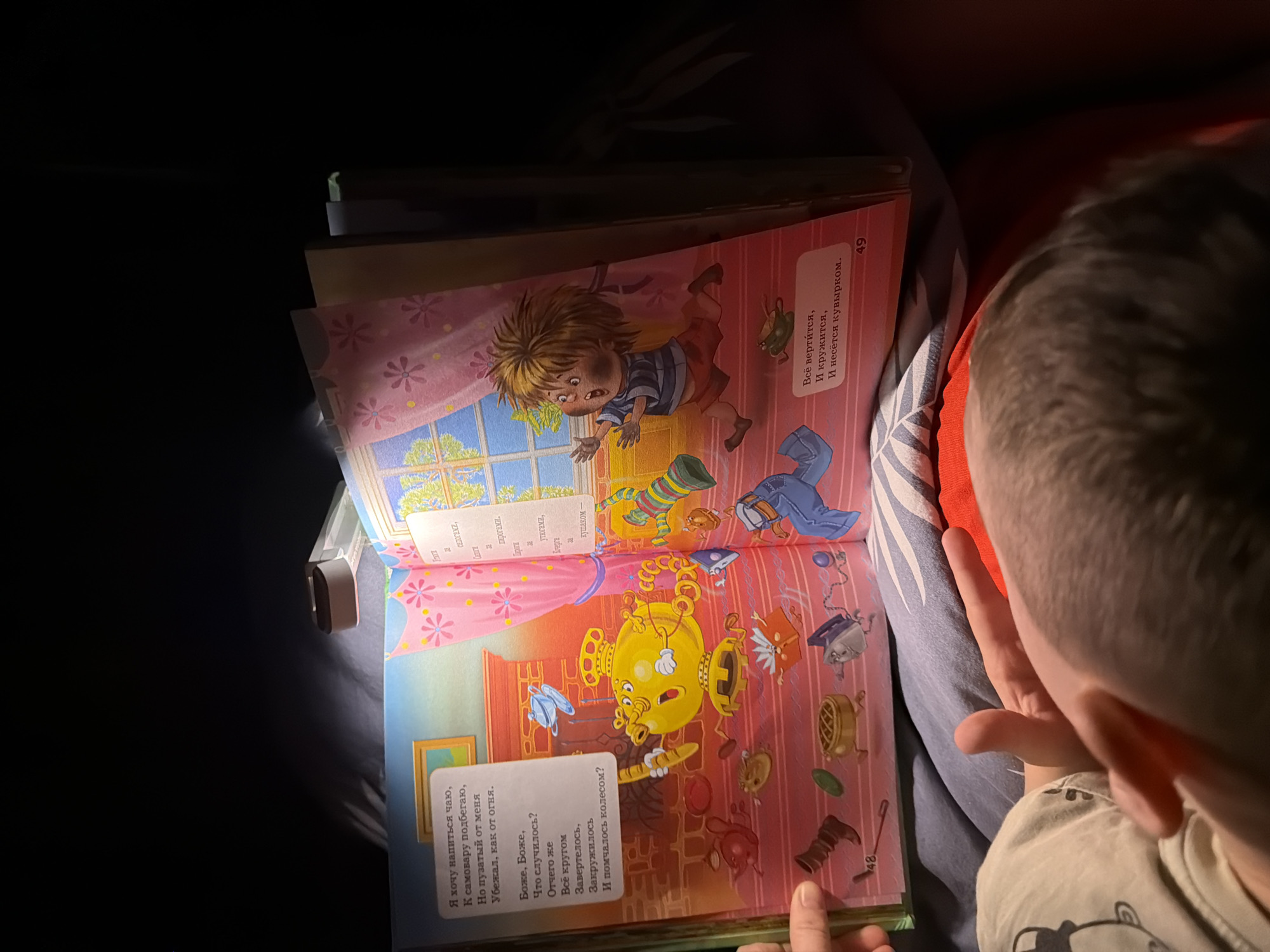 The image shows an open children's book illuminated by a portable LED light, casting shadows. The book features colorful illustrations and Russian text. On the left page, a text box reads: "Я хочу напиться чаю, К самовару подбегаю, Но пузатый от меня Убежал, как от огня. Боже, Боже, Что случилось? Отчего же Всё кругом Завертелось, Закружилось И помчалось колесом?" Below this text is an illustration of a runaway, anthropomorphic samovar with a panicked expression, rolling away from a room that appears to be a living space with a framed picture on the wall and a brick-patterned wall. Scattered on the floor are various objects such as a boot, a flattened tin can, a pie, and an iron. The number "48" is visible at the bottom left of this page.
The right page of the book depicts a young boy with messy blond hair running away, his arms outstretched, with a look of alarm on his face. He is wearing a striped shirt and red shorts. A pink curtain with flower patterns hangs to the left of the boy, framing a window with a view of trees. Another text box on this page reads: "Всё вертится, И кружится, И несётся кувырком." The number "49" is visible at the bottom right of this page.
The book is resting on a bed covered with a blue and white patterned comforter and a red blanket. A person's hand is visible on the bottom left corner of the image, holding the edge of the book. The overall scene suggests a person is reading or looking at the book in a dimly lit environment. The image was captured in Khabarovsk, Russia.