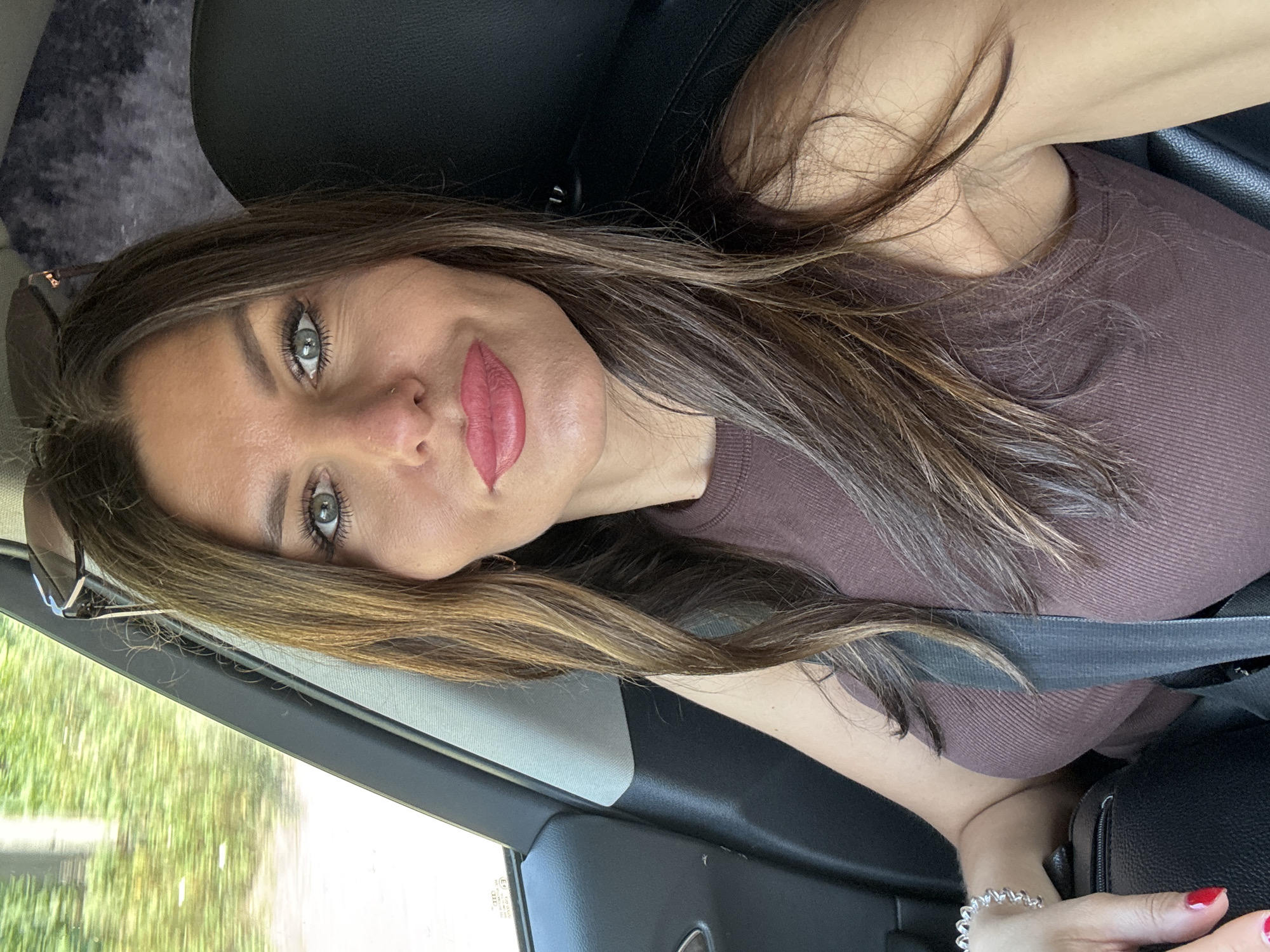 A woman with long, brown hair and green eyes, wearing a brown tank top, is sitting in the passenger seat of a car. She is wearing a seatbelt, a silver bracelet, and red nail polish. Her makeup includes lipstick and eyeliner. Sunglasses are perched on her head. The background shows trees and foliage through the car window, suggesting a location outside of a city, possibly in a rural or suburban area. The specific location within Noviy, Russia cannot be determined from the image.