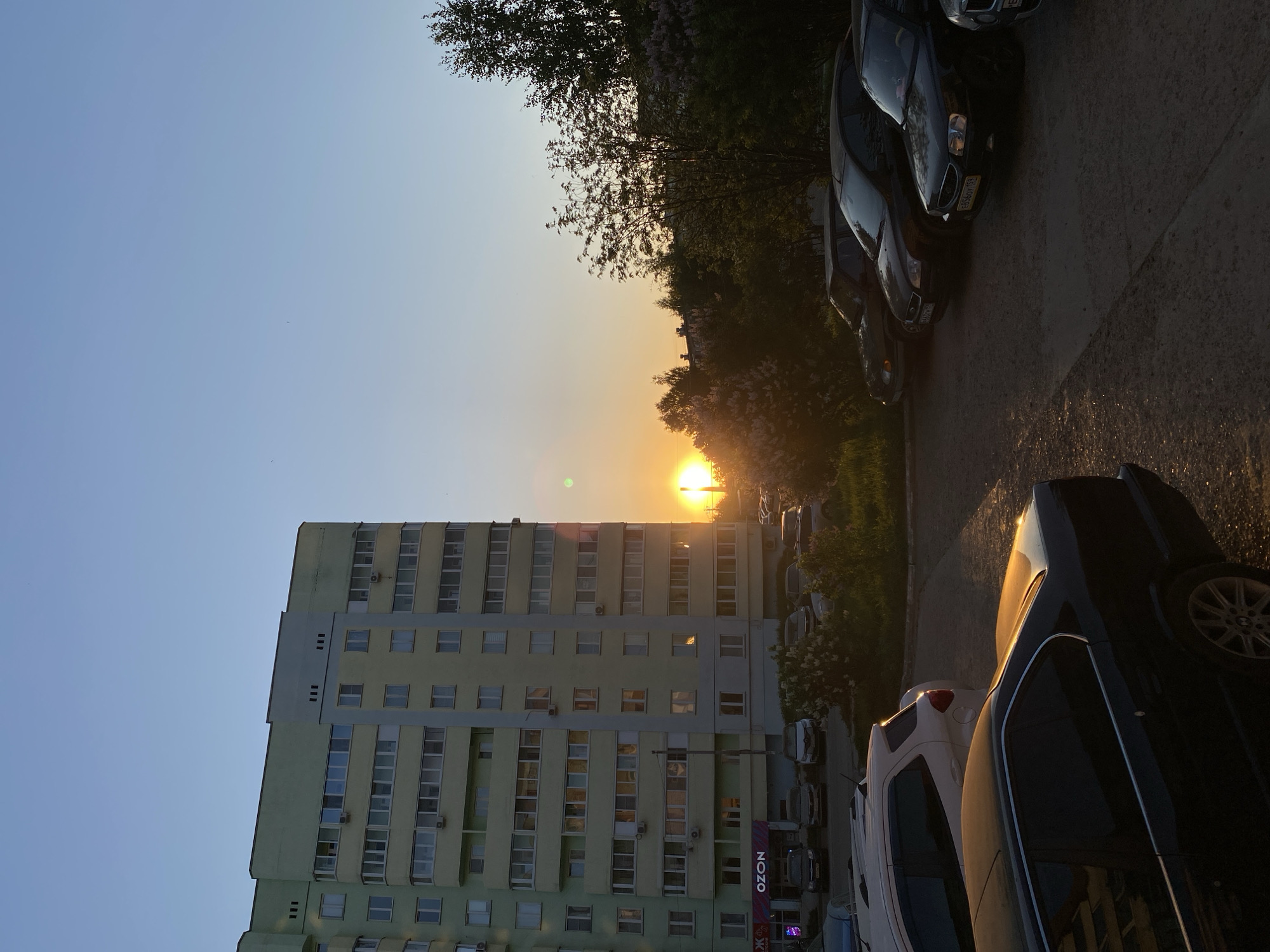 The image shows an urban setting in Chaikovsky, Russia, during sunset. The foreground features several parked cars lining the side of a road. A large apartment building is prominent on the left side of the frame. The setting sun is positioned in the center of the image, partially obscured by trees and other buildings in the distance. The sky transitions from blue at the top to orange near the horizon where the sun is setting. An "OZON" sign is visible on the lower right side of the apartment building.