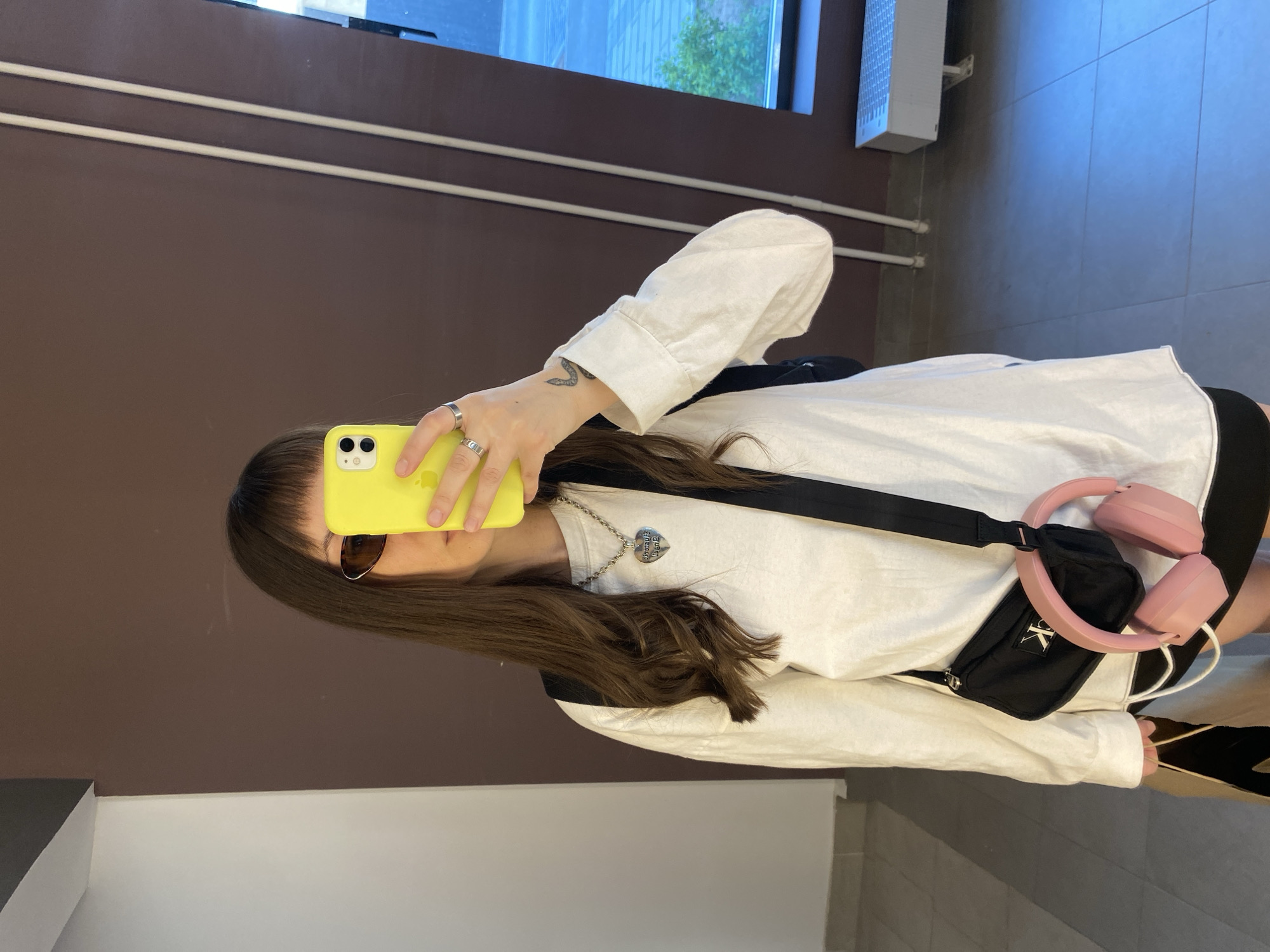 A person with long brown hair, sunglasses, and a white long-sleeved shirt is holding a yellow phone up in front of their face, seemingly taking a selfie in front of a mirror. The person has a black fanny pack with pink headphones attached and a silver necklace with a heart pendant. The background consists of a brown wall with white trim, a window with a view of greenery, a blue and white tiled floor, and a white lower wall. The location is likely Izhevsk, Russia, given the image's origin.