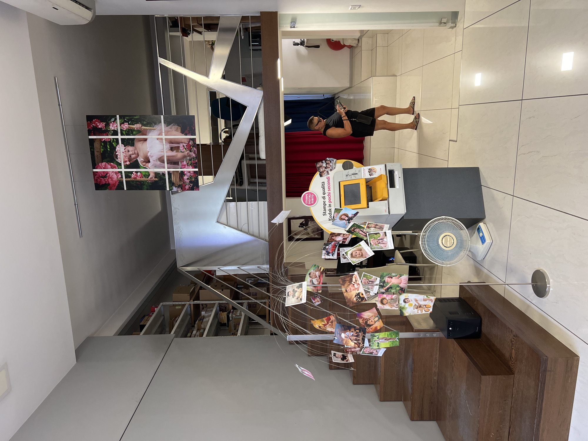 The image depicts the interior of a photography studio or a similar establishment, likely in Bitonto, Italy. The scene is characterized by a modern staircase with metal railings, leading to an upper level. A prominent display of framed photographs hangs on a wall near the staircase, showcasing images of children, including one of a baby on a swing surrounded by flowers.
In the foreground, a spiral mobile made of metal wires holds numerous small photographs, predominantly featuring families and children, creating a whimsical display. To the right, a yellow Kodak photo printing station is visible, suggesting the service offered at this location. A woman in a black dress and sandals stands near the photo station, looking at her phone.
Other notable objects include a white oscillating fan, a black UPS unit, and shelving units filled with boxes in the background. The lighting suggests it is daytime, with artificial lights also contributing to the illumination. The overall atmosphere appears to be that of a commercial space focused on photography services. Text visible on a circular sign near the photo station reads "Stampe di qualità Kodak in pochi secondi" (Quality Kodak prints in seconds).