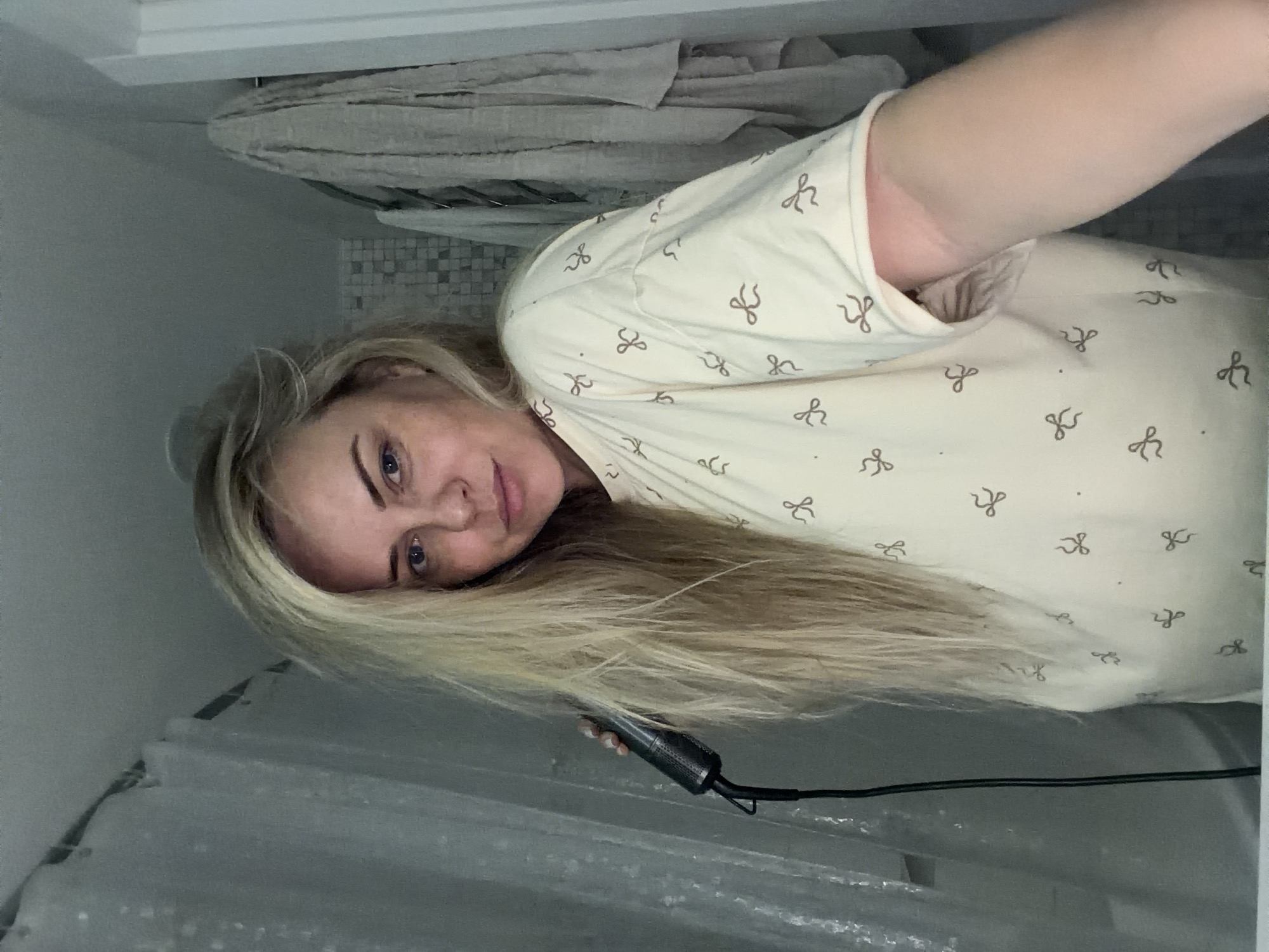 The image, oriented vertically on its side, shows a woman's upper body and face in what appears to be a bathroom. The woman has long, straight blonde hair that covers part of her chest and extends towards the bottom of the frame. Her face is turned towards the viewer, revealing blue eyes and a neutral expression. She is wearing a light-colored garment, possibly a t-shirt or pajama top, patterned with small, dark, bow-like shapes.
In her right hand, she holds a black hair styling tool, likely a curling iron or straightener, with a textured handle and a black cord extending from it. The activity appears to be hair styling, or a moment captured during the process.
The background elements indicate an indoor setting, specifically a bathroom. In the foreground, a translucent shower curtain with a slight texture is visible. Behind the woman, there's a wall with a section of small, grey and white mosaic tiles. Above this, folded light-colored towels are stacked on a shelf or rack. A plain white shelf or ledge runs along the top edge of the background. No visible text is present in the image. The time of day and weather are not discernible.