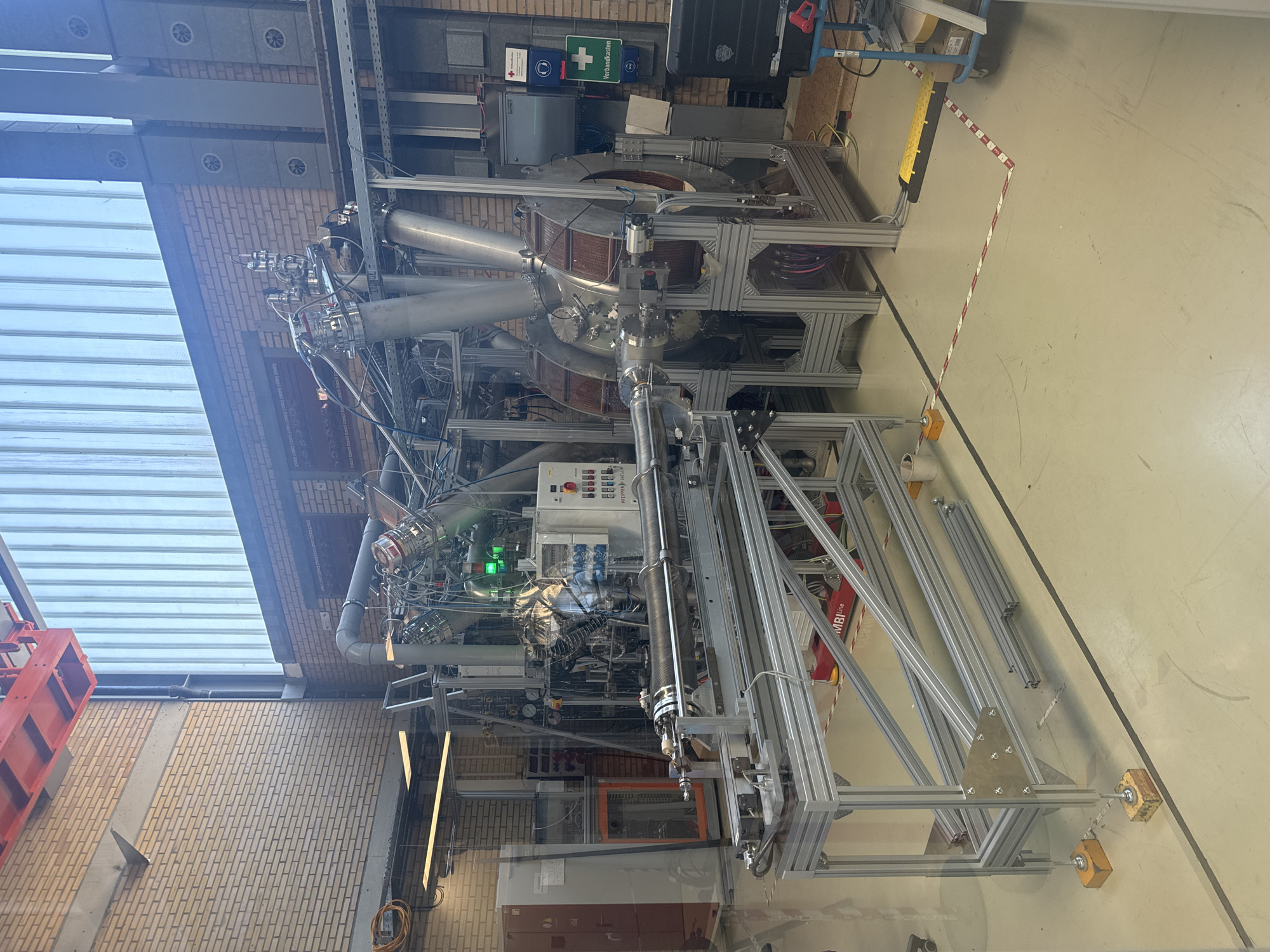 The image shows a laboratory at the Forschungszentrum Jülich in Jülich, Germany. The laboratory contains a large, complex scientific apparatus, likely for experimental physics or engineering research. The apparatus consists of various metallic components, including pipes, vacuum chambers, coils, and a control panel. It is mounted on an aluminum frame structure. In the background, a brick wall with large windows and industrial ventilation systems is visible. There are also various pieces of laboratory equipment, such as a large cabinet on the left and a rolling cart with equipment on the right. The floor is a light-colored concrete with black lines and yellow safety markings. There are no people visible in the image. The scene is well-lit, likely from overhead laboratory lighting and natural light from the windows.