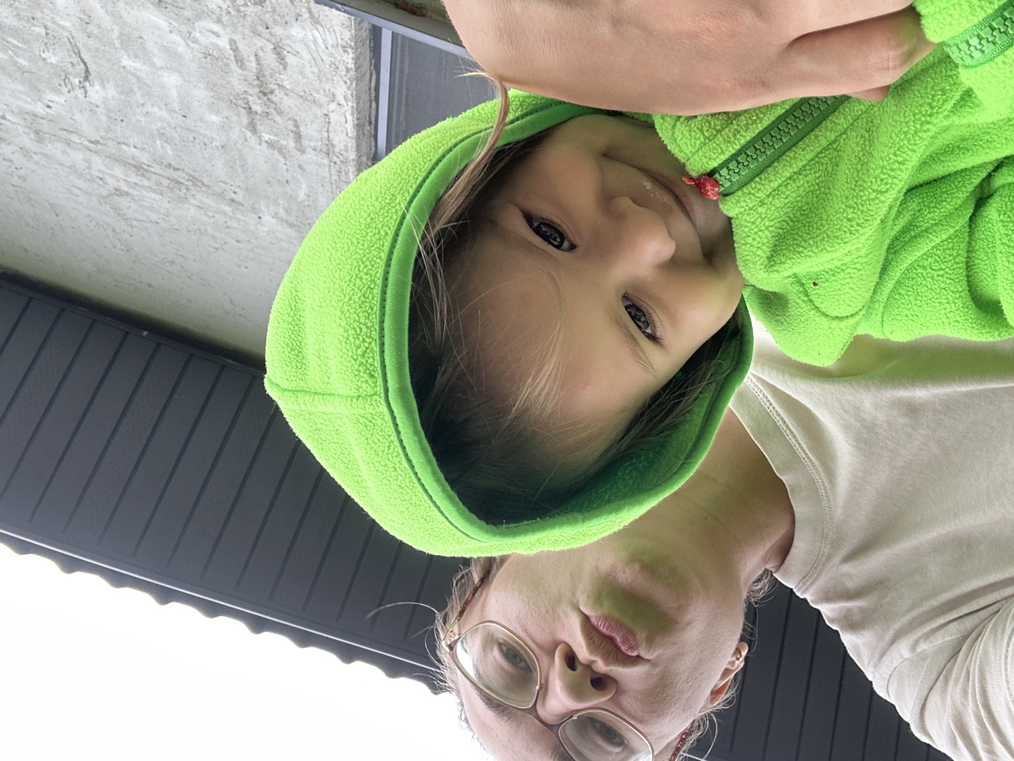 A low-angle, close-up selfie shows a woman and a young child outdoors. The woman, wearing glasses and a light-colored t-shirt, makes a puckered face towards the camera. The child, wearing a bright green hooded jacket, smiles and leans into the frame. The background consists of a dark, ribbed roof structure and a textured, light-colored wall, suggesting a residential setting. The scene appears to be in the city of Kharnavy, Russia.