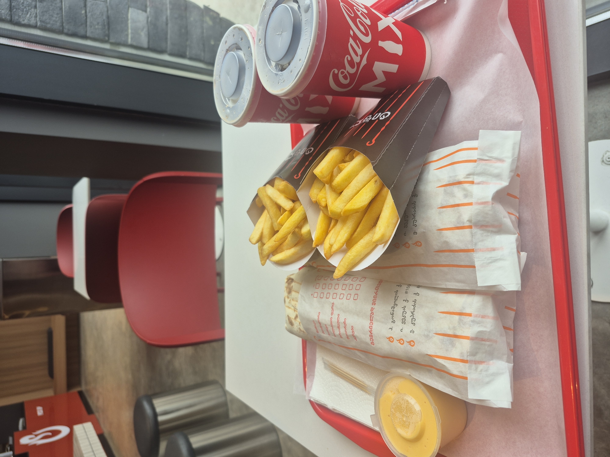The image shows a red tray of fast food, presumably in Tbilisi, Georgia. Two red Coca-Cola Mix cups with lids stand at the top of the tray. Below them are two brown cartons of french fries, with yellow fries visible. Two wraps wrapped in white paper with orange stripes and Georgian text are stacked below the fries. At the bottom right of the tray is a small clear plastic container filled with a yellow sauce. The tray sits on a white surface, likely a table, near a window. In the background are red chairs and gray steel legs of other chairs or tables. The Georgian text on the wraps references addresses, including "I. Vekua st. #159," "V. Kupradze st. #," and "G. Gonashvili st." and names of ingredients: "pomegranate," "mayonnaise," and "ketchup."