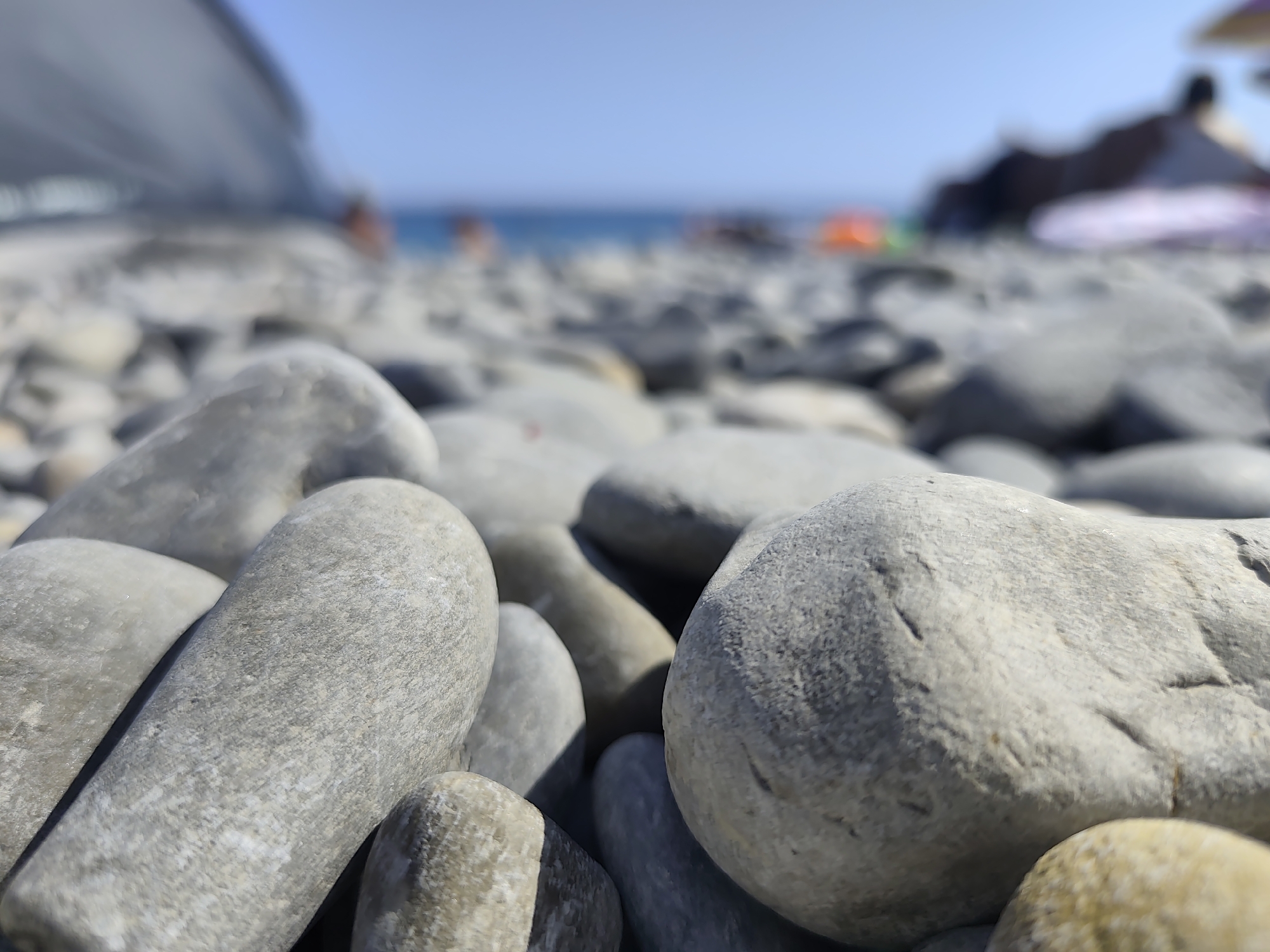 This is a close-up, low-angle photograph of a pebble beach, likely in Nice, France, given the context. The foreground is dominated by smooth, gray and white pebbles of varying sizes, some appearing elongated and others more rounded. The pebbles are arranged densely, creating a textured surface. In the mid-ground, the pebbles continue, but the focus softens, and the shapes become less distinct. Beyond the beach, the blue sea is visible under a clear, pale blue sky. The horizon line separates the sea from the sky. In the far distance, blurred figures of people can be seen, along with what appears to be beach umbrellas and structures, suggesting a populated beach environment. The overall lighting indicates a bright, sunny day.