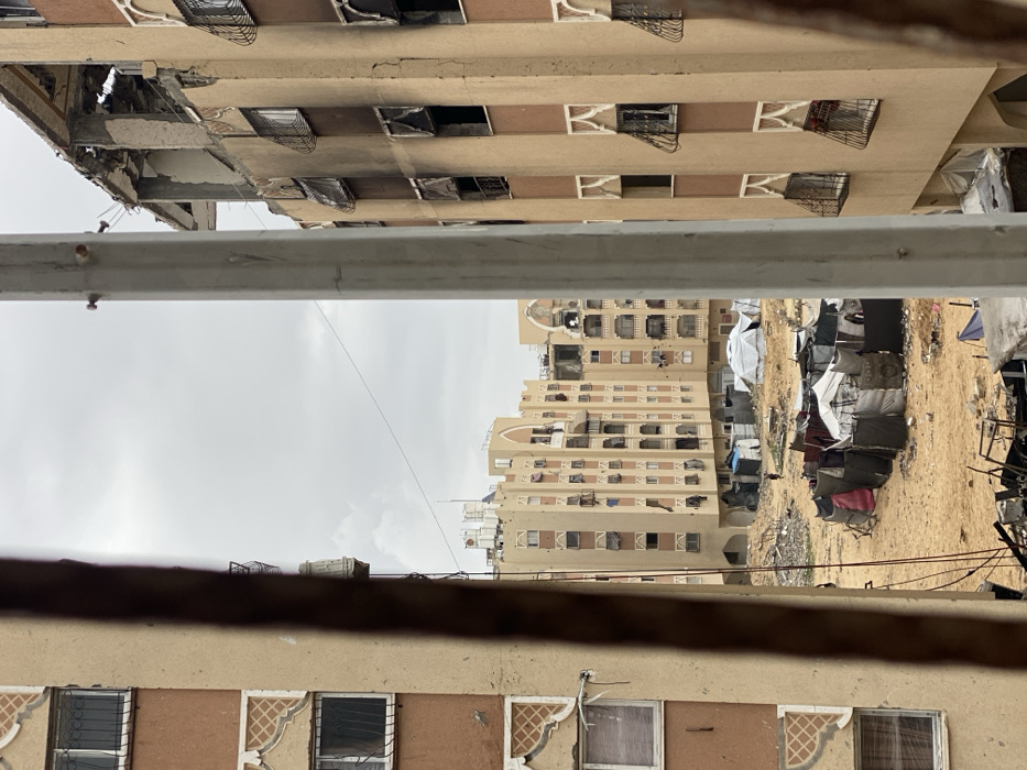The image displays a panorama of a heavily damaged urban residential area in Khan Yunis, Palestinian Territory, viewed through two vertical structural elements. On the right, a multi-story building features a light tan facade with vertical brown strips; its windows, many equipped with decorative white arches and wrought iron grates, are largely blackened by fire. The upper floors show significant structural destruction, including collapsed sections and exposed rebar. A similar building is partially visible on the left. The central ground area between the buildings is an expansive, debris-strewn dirt field containing numerous improvised shelters constructed from tarps, fabric, and miscellaneous materials. Further back, at least one larger, white tent is visible among the shelters. A lone, indistinct figure is walking in the sandy area in the mid-distance. In the background, multiple other multi-story residential buildings, sharing similar architectural styles, exhibit varying degrees of damage under an overcast sky.