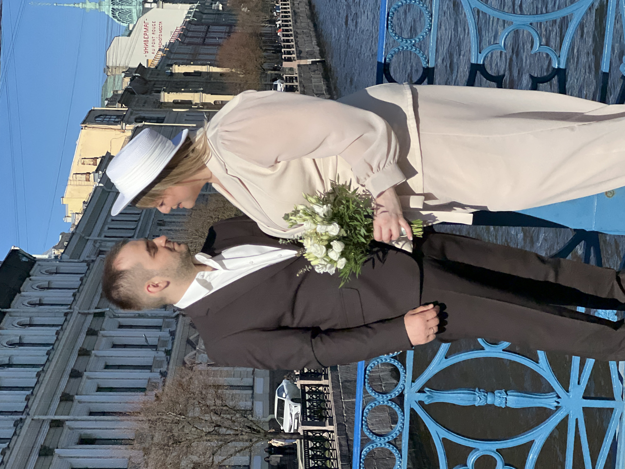 A newlywed couple is posing on a blue metal bridge in Saint Petersburg, Russia. The woman is wearing a light pink dress and a white hat and holding a bouquet of white flowers. The man is wearing a dark suit and a white shirt. They are facing each other with their heads close together. Buildings and a waterway are visible in the background. The sign "УНИВЕРМАГ- AU PONT ROUGE ПЕРВОГО МОСТА" is visible on one of the buildings.