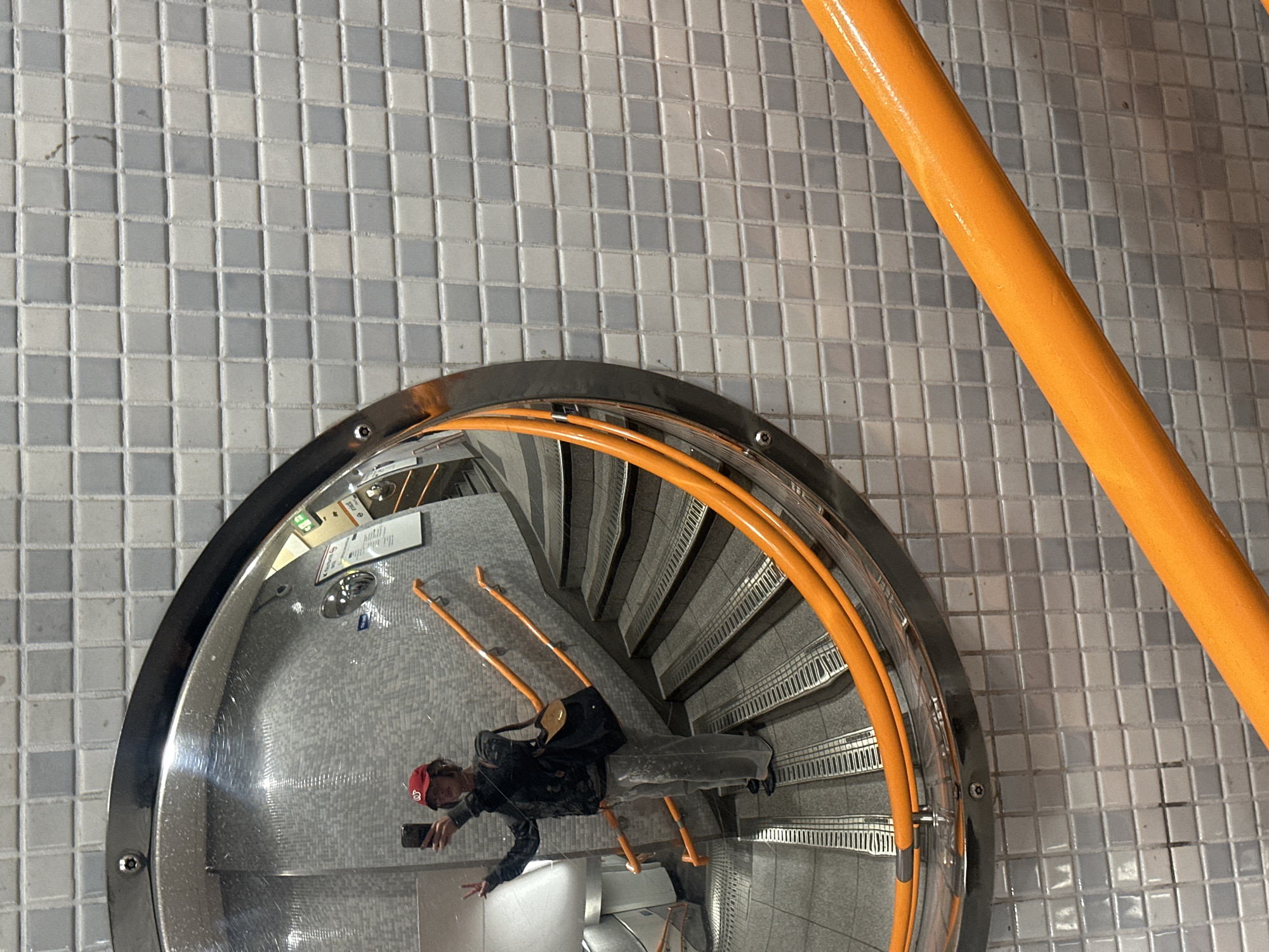 A high-angle, distorted view captures a man taking a photo of himself in a circular mirror mounted on a wall tiled with small grey and white squares. The man, wearing a red baseball cap, dark jacket, and trousers, holds a smartphone in his left hand and extends his right hand. The mirror reflects a spiral staircase with orange handrails. A portion of an orange vertical support beam is visible in the upper right corner. The image is potentially taken in the London Underground, specifically the curved walls of the escalators in underground train stations.