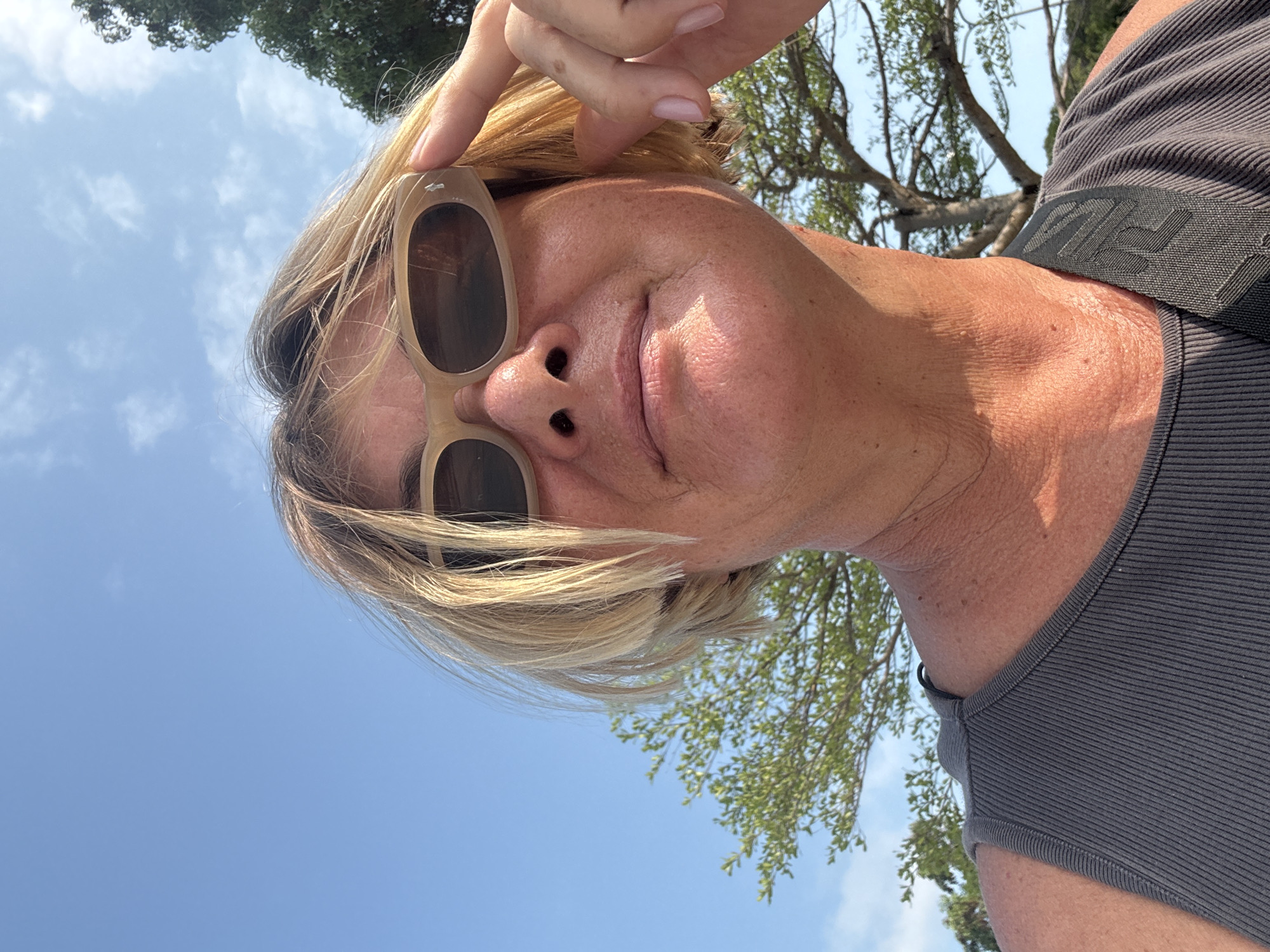A low-angle shot shows a light-skinned person with short blonde hair and sunglasses pushed slightly down the nose. The person is wearing a dark grey ribbed sleeveless top with a black strap visible on the right shoulder. The background includes a bright blue sky, tree branches, and foliage. The location cannot be verified as บ้านบางรักษ์, Thailand.
