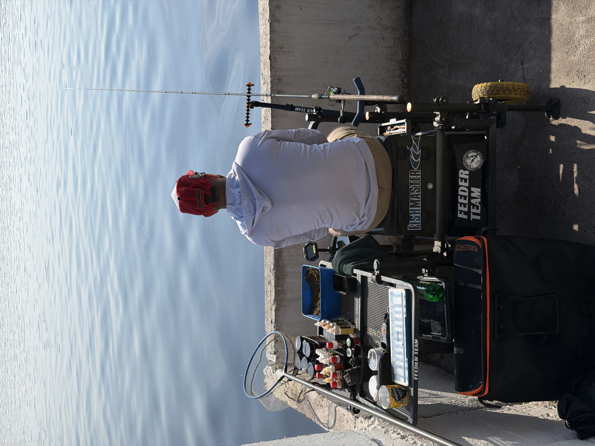 The image depicts a person fishing on the bank of a body of water, likely the Kama River, in Chaikovsky, Russia. The individual, facing away from the camera, is seated on a fishing station and is wearing a red baseball cap and a light-colored hoodie. To their right, a fishing rod is set up in a rod holder. The fishing station is equipped with various tackle, including a net, bottles of attractants, and a tackle box. A large black bag with orange trim is positioned below the station. The water is calm with gentle ripples, and the sky is clear, suggesting fair weather. The overall scene conveys a moment of quiet leisure and outdoor activity.