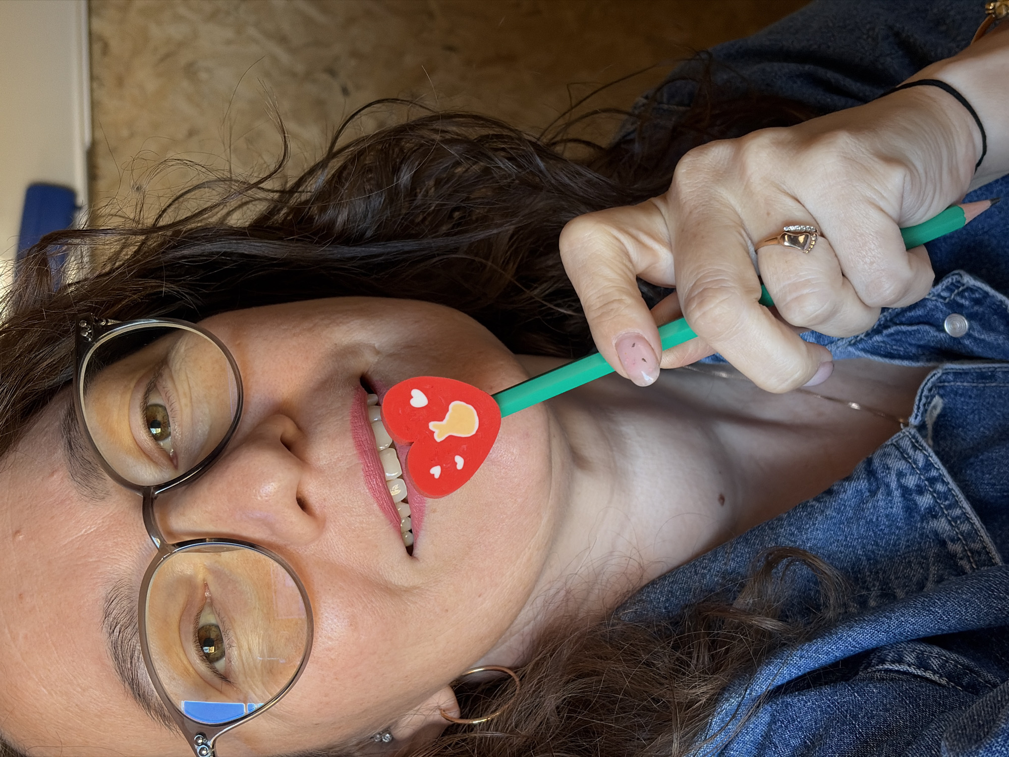 This is a close-up, eye-level, outdoor, waist-up shot of a woman in her 40s, wearing glasses and a denim jacket. The woman has brown hair and brown eyes. She is holding a green pencil with a heart-shaped eraser in front of her mouth. The heart eraser is red with small white hearts and a yellow hand gesture. Her lips are painted pink. She is wearing a gold ring with diamonds on her right ring finger. The background is blurry and appears to be indoors, with a wooden wall visible on the right. There is no indication of the city of Ol'khovskoye, Russia.