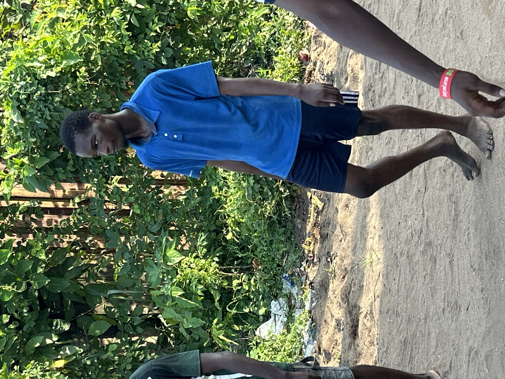 A full-body shot of an African man wearing a blue polo shirt and dark shorts is standing in Ihiagwa, Nigeria. He is looking downwards. He is barefoot, and his feet are soiled. Another person is partially visible with a red wristband. The background is foliage and a weathered wooden fence. There is debris on the ground. Part of another person is visible on the lower left, wearing a green shirt. The ground is sandy and dusty.