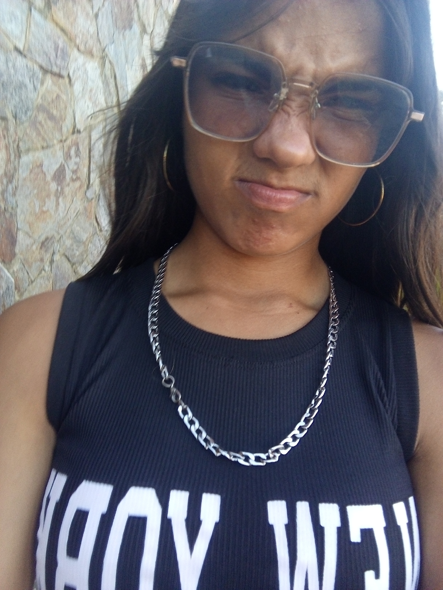 A woman poses for a selfie in front of a stone wall. She is wearing sunglasses, hoop earrings, and a silver chain necklace. Her black tank top has large white letters on the front. Her facial expression is a scowl. The location is identified as Juan Griego, Venezuela.