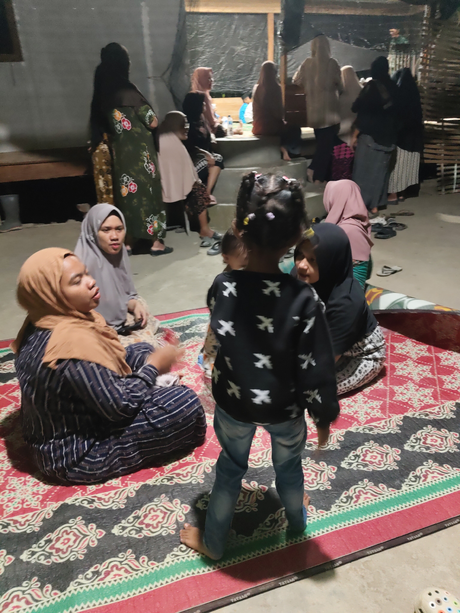 In Gampongbaro, Indonesia, a group of people are gathered in what appears to be a communal area. In the foreground, two women are seated on a large, patterned mat. One woman wears a light brown hijab and a striped dark blue and white garment, and is gesturing with her hands. The other woman wears a light grey hijab and a patterned skirt. Behind them, a young child with braided hair, wearing a black sweatshirt with white airplane patterns and blue jeans, stands facing away from the camera. Several other individuals, mostly women wearing hijabs and modest clothing, are visible in the background, some standing and others seated. The environment seems to be an open-air structure with concrete flooring and wooden elements. The lighting is dim, suggesting it might be evening.