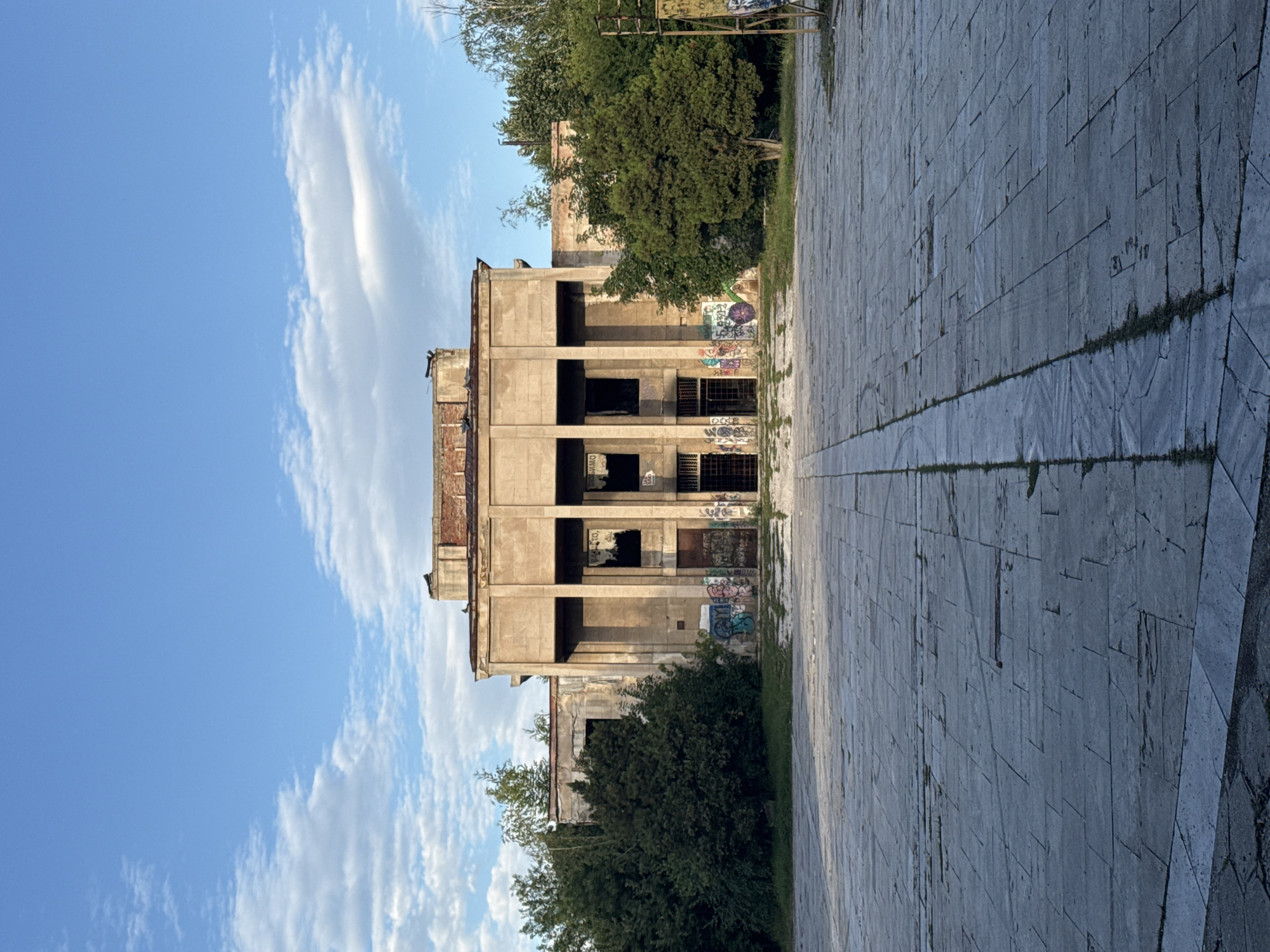 The image depicts the dilapidated facade of the former Palace of Culture in Pernik, Bulgaria. The structure is characterized by its imposing concrete columns and large, dark, empty window openings, many of which are boarded up or covered with graffiti. The building appears abandoned and is in a state of decay, with exposed brickwork on the upper levels and crumbling concrete. Dense green foliage surrounds the building, encroaching on its grounds. In front of the structure is a wide, paved plaza, its stone slabs cracked and worn, with patches of grass growing through the gaps. The sky above is a clear blue, dotted with scattered white clouds, suggesting it is either morning or late afternoon. The overall scene conveys a sense of neglect and the passage of time, with the once grand building now a subject of urban decay. There are no people visible in the image.