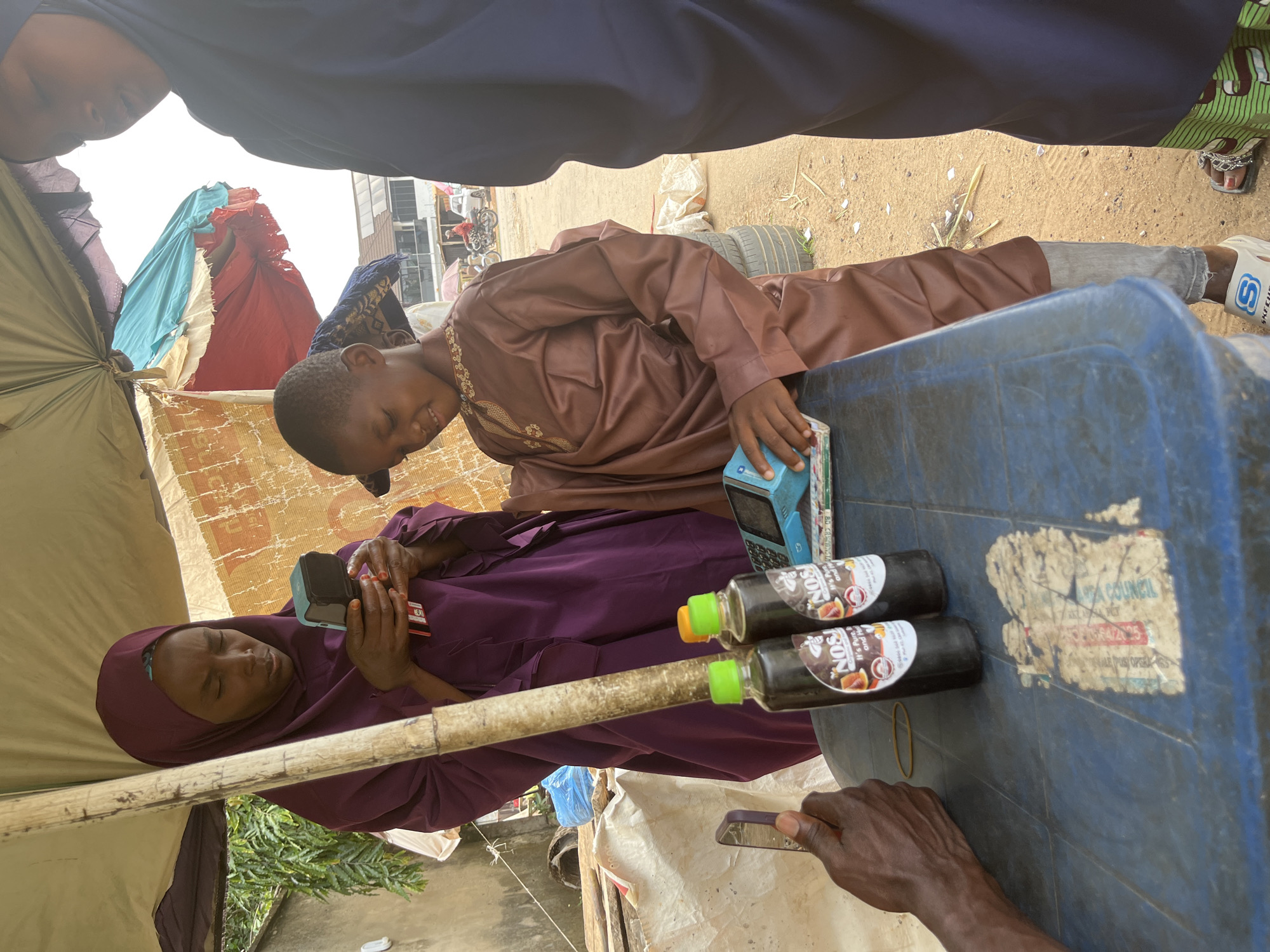 This media file shows a group of people, likely a family, under a makeshift canopy. A young boy, dressed in a brown kaftan, smiles and looks towards a tablet device, possibly playing a game. Beside him, a woman in a purple hijab is also looking at a phone, with a cash register and two bottles of what appears to be juice or a health drink on a blue table in front of her. The scene is set outdoors, with sandy ground and some greenery visible. The light suggests it is daytime. The overall atmosphere seems relaxed and casual. There are some torn fabrics and blankets that make up the canopy. On the bottles, the visible text reads "NOS. It's Pure and Natural." On the cash register, there are some indecipherable labels. The scene likely takes place in a market or roadside stall setting in Abuja, Nigeria.