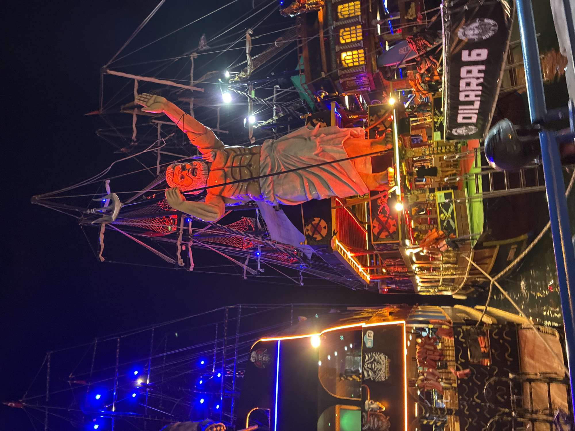 The image shows two pirate-themed boats docked side-by-side at night in Alanya, Turkey. The boat on the right is larger and features a prominent statue of a muscular, bearded man in a draped garment at its bow, with his arms raised. This boat is illuminated with red and yellow lights, and has elaborate decorations on its railings and windows. A banner reading "DILARA 6" is visible on its side. The boat on the left is also decorated with lights, including blue and orange neon strips, and displays skull motifs. The water between the boats reflects the colorful lights. The overall scene suggests a lively nighttime tourist attraction.